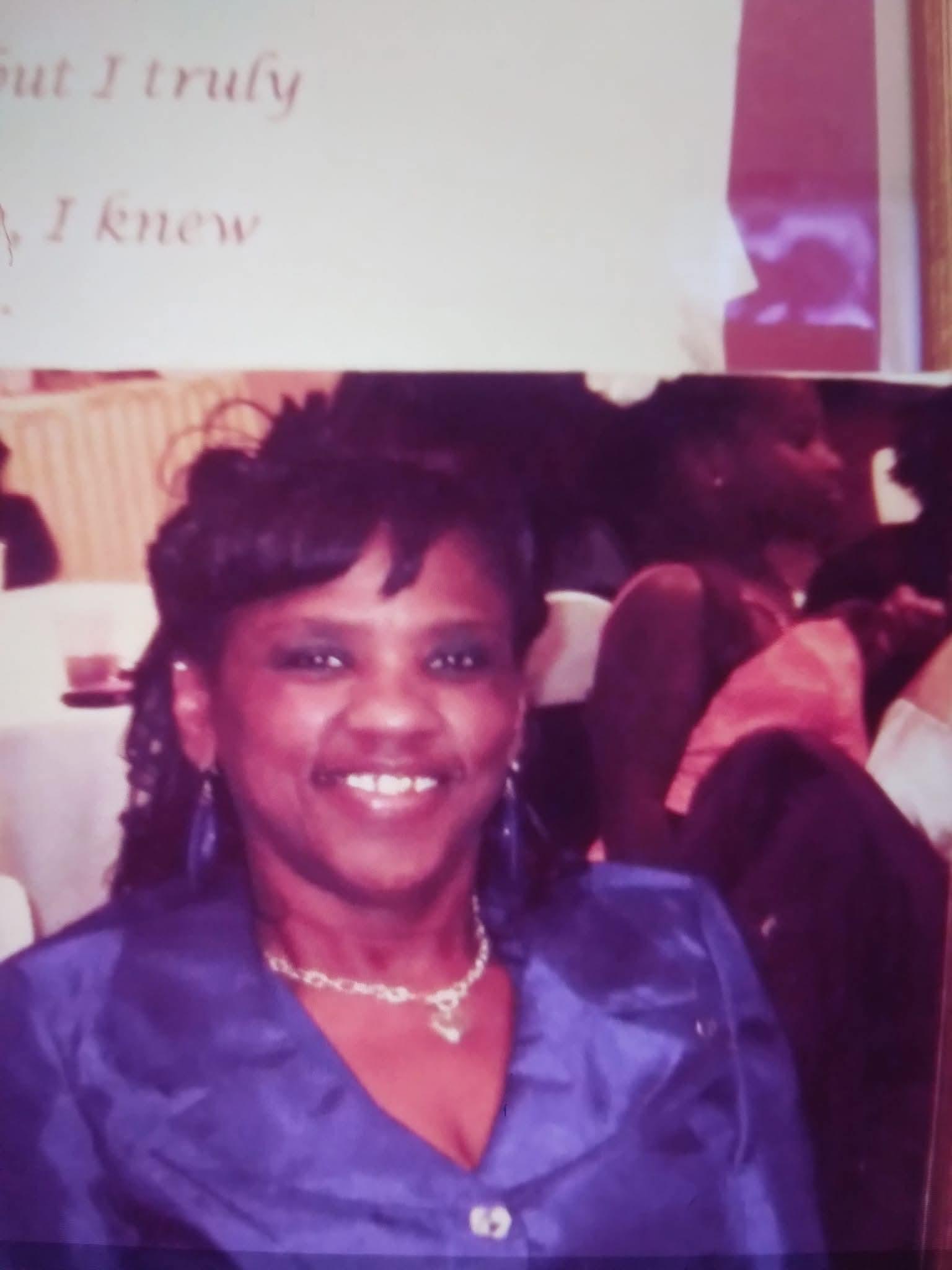 A joyful woman wearing a blue outfit smiles warmly at a gathering filled with guests.