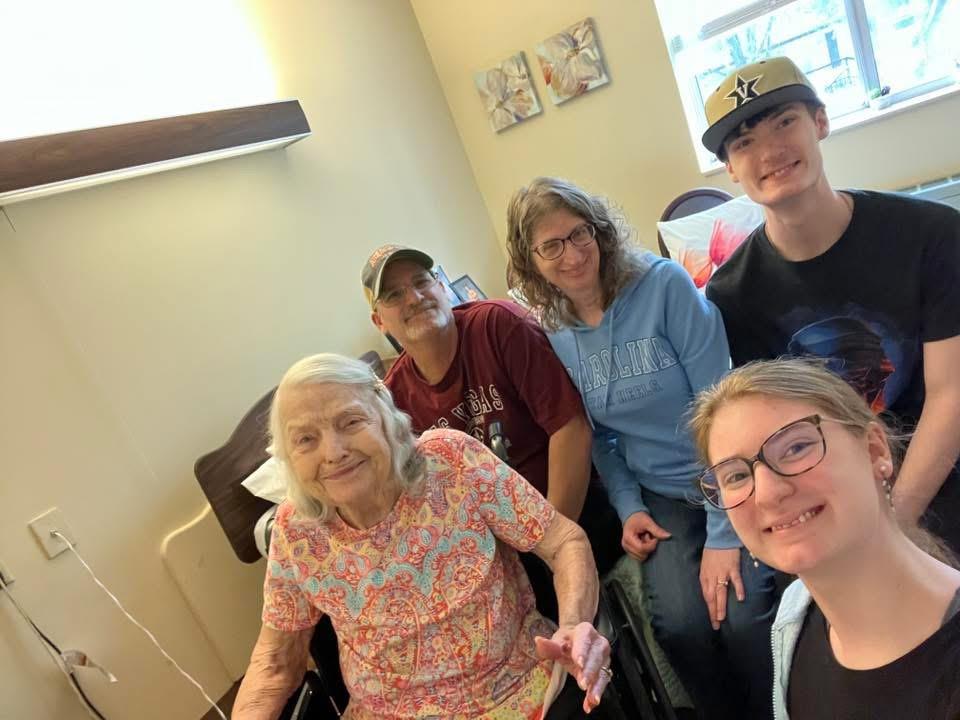 Family members smile warmly while visiting their loved one in a care facility room.
