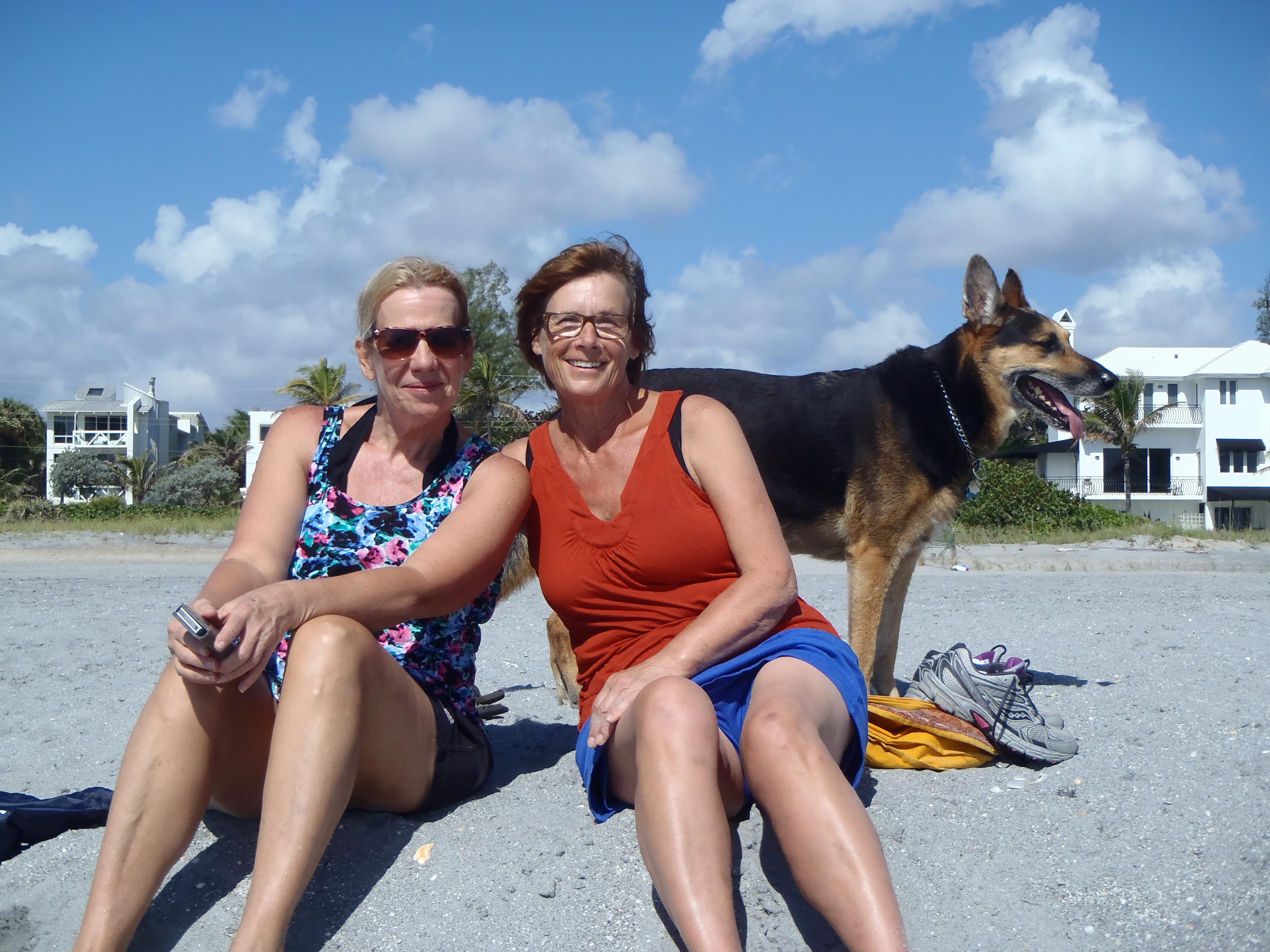 Two women sit together on a sandy beach, smiling and enjoying the sunshine with a dog nearby.