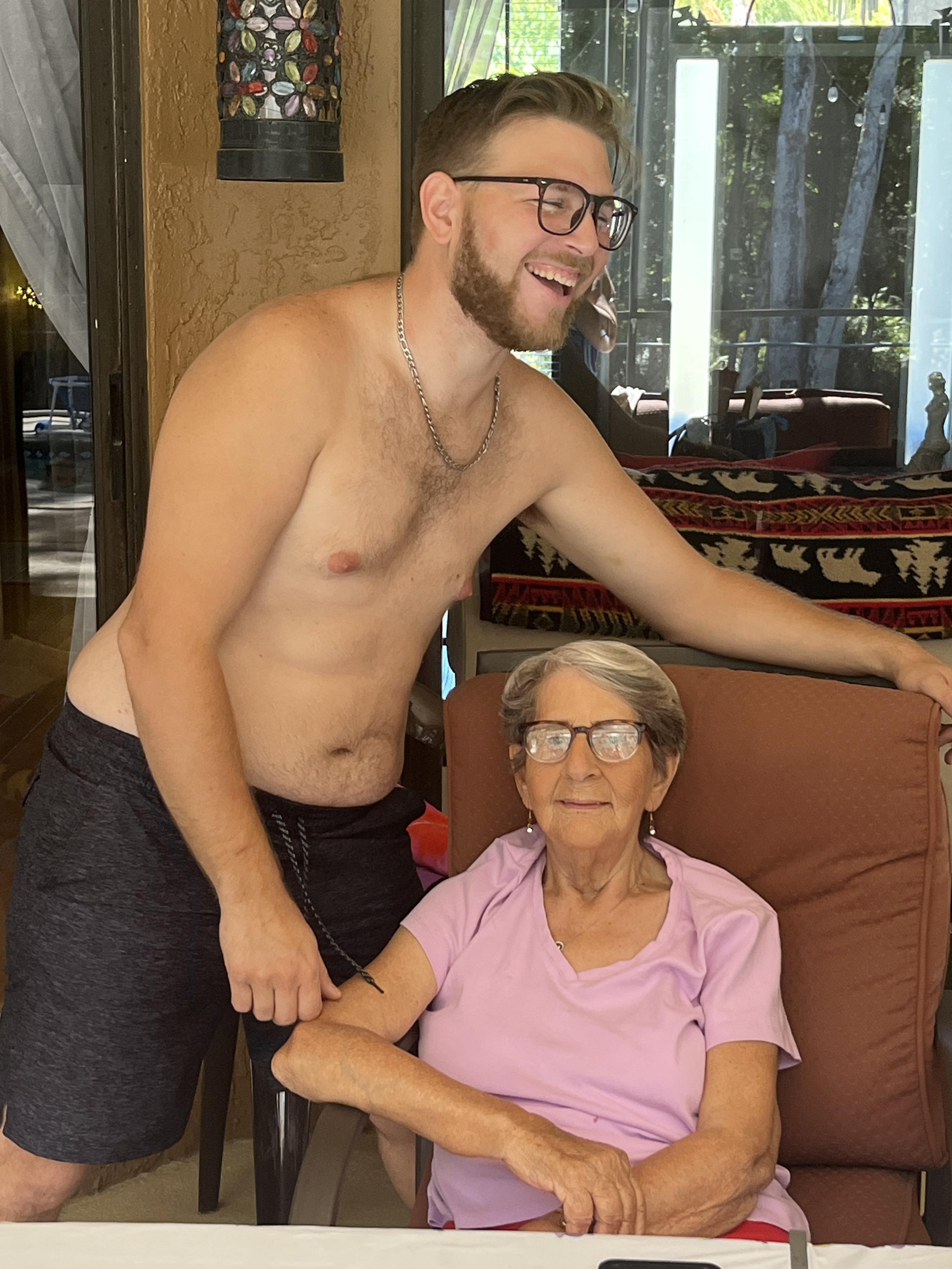 A young man playfully stands next to his grandmother, both smiling in a relaxed outdoor setting.