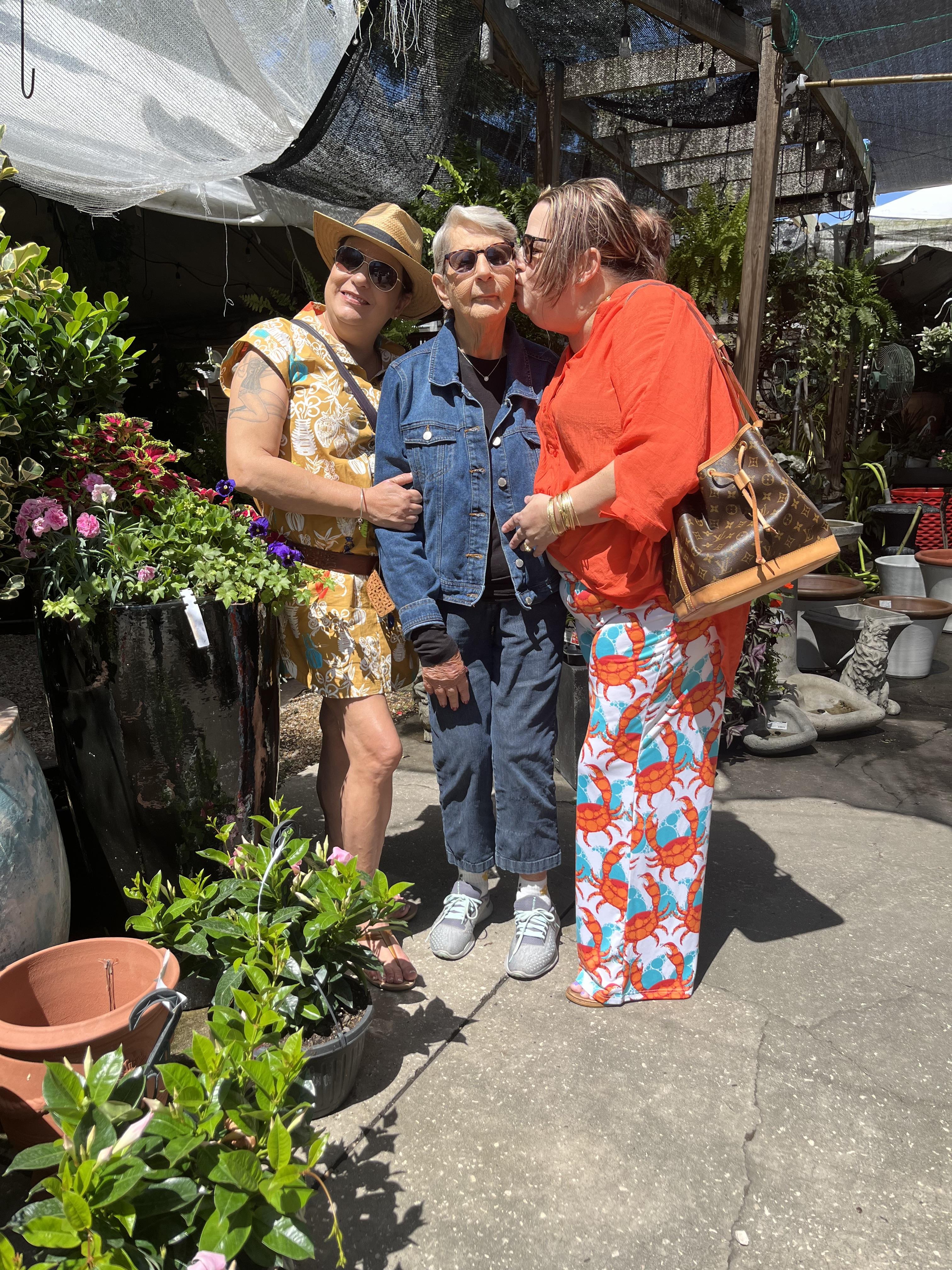 Three friends smile together at a vibrant flower market, enjoying the day among plants.