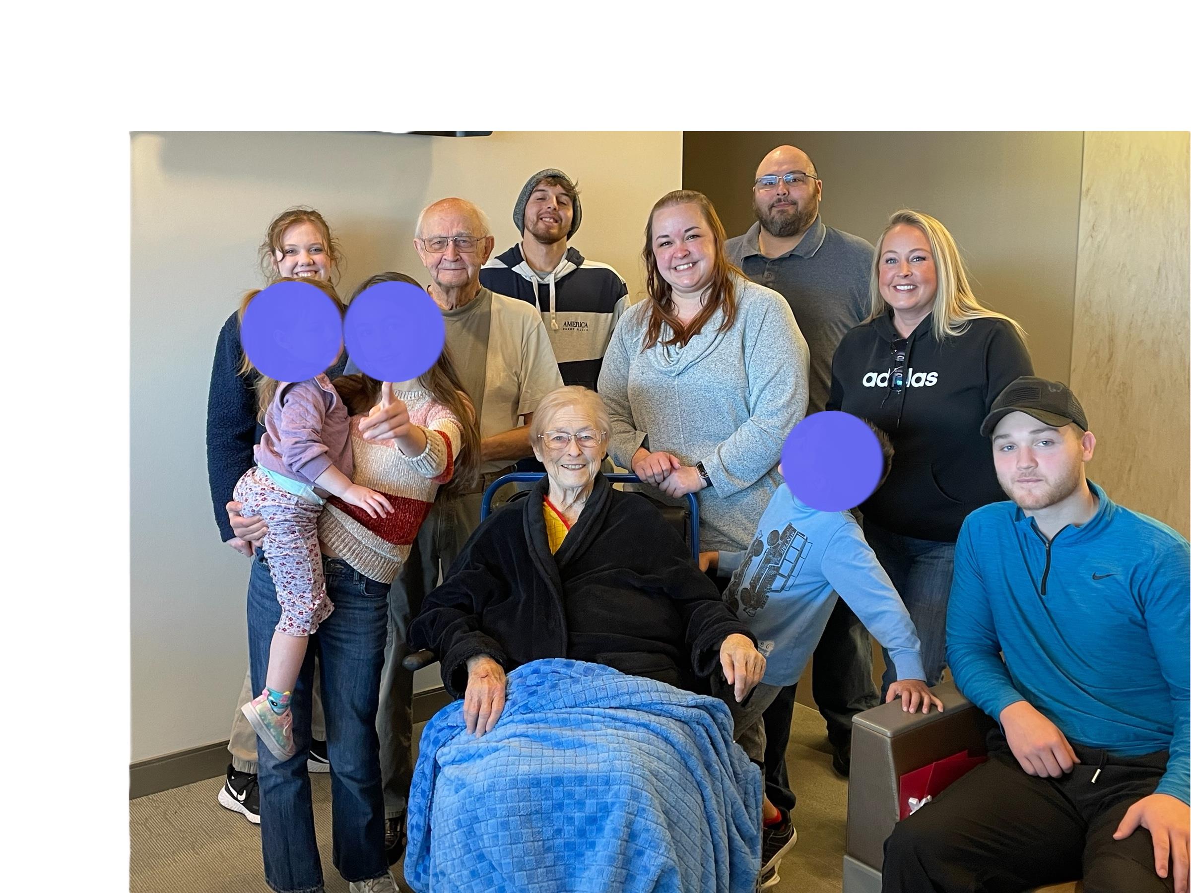 Group of family members poses happily with an elderly woman in a care facility during a visit.