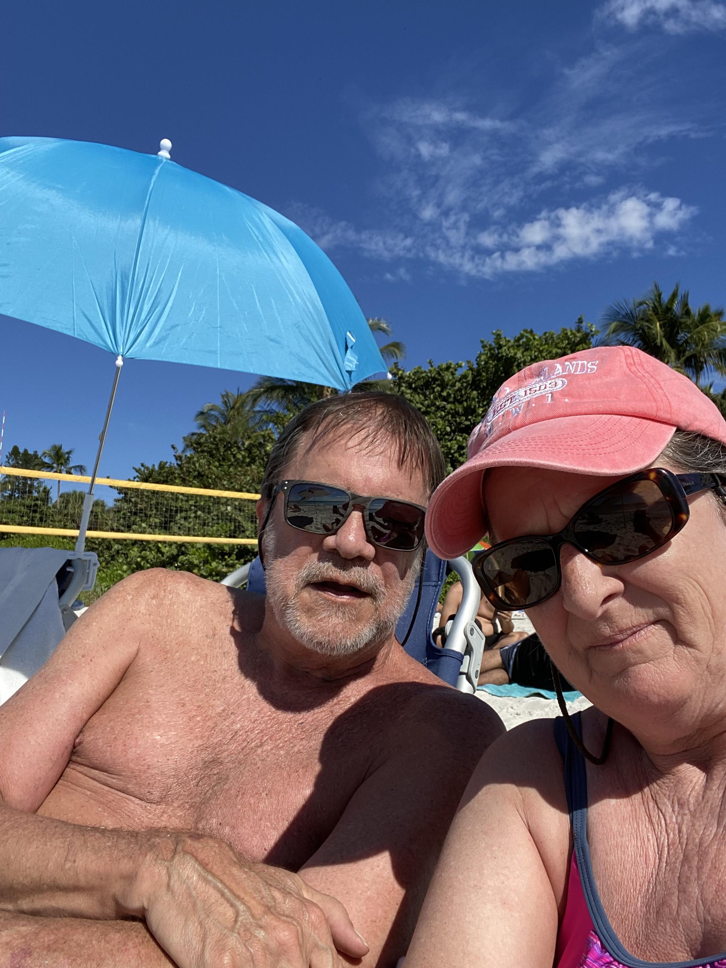 Two people relaxing on the beach, wearing sunglasses and enjoying the sun beneath a blue umbrella.