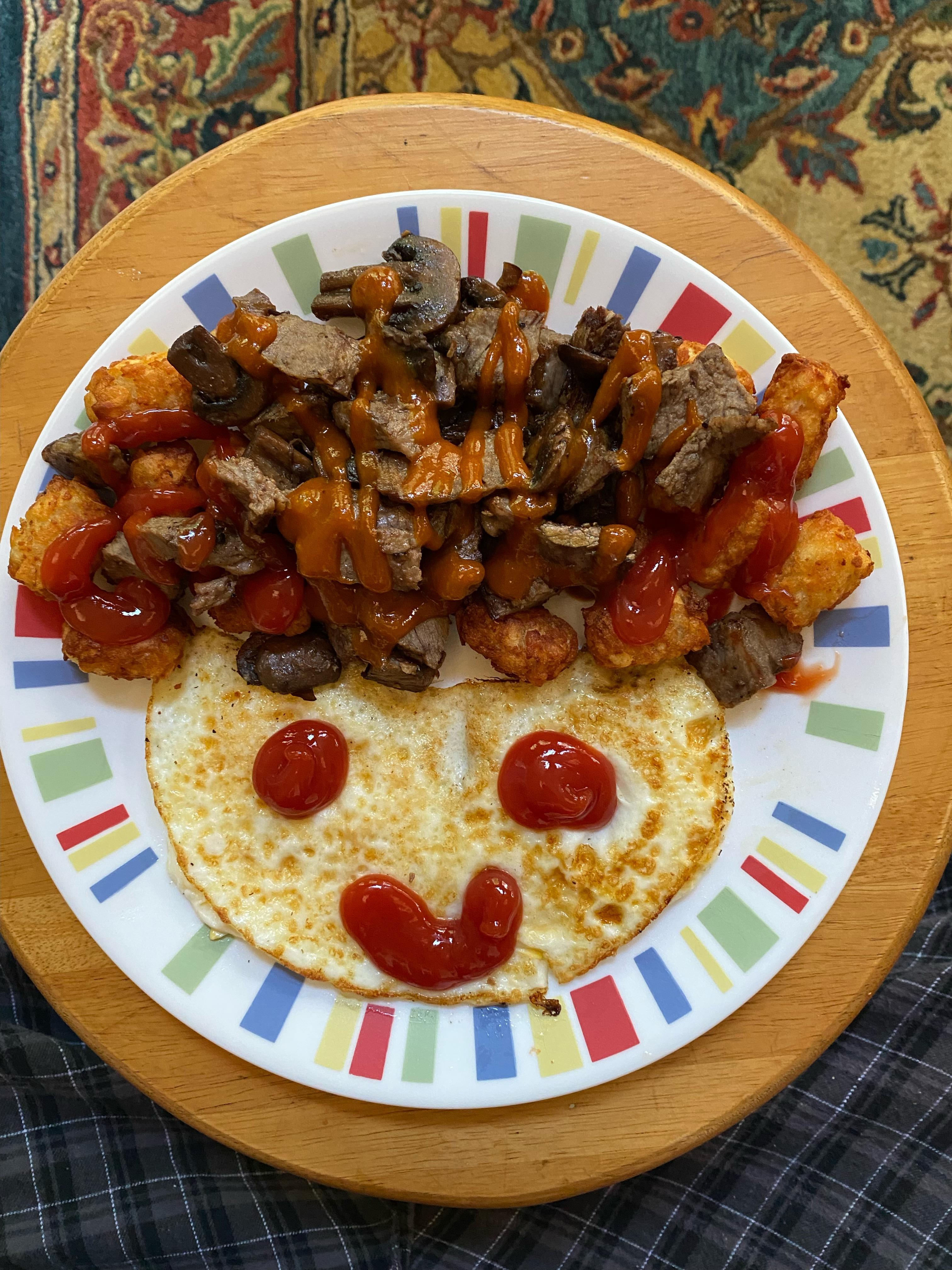 Delicious breakfast with a smiley face of eggs, ketchup, steak, and tater tots.