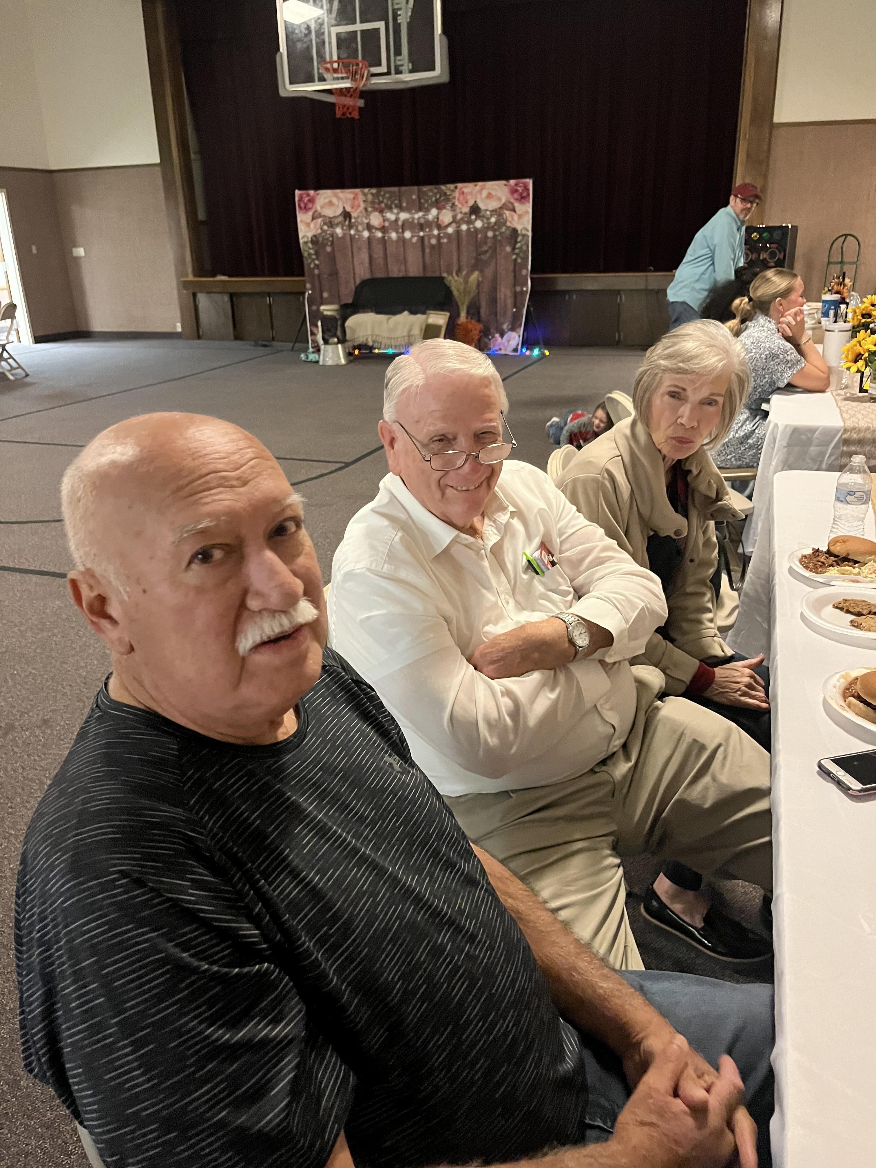 Seniors gather at a long table, chatting and enjoying food in a community hall.