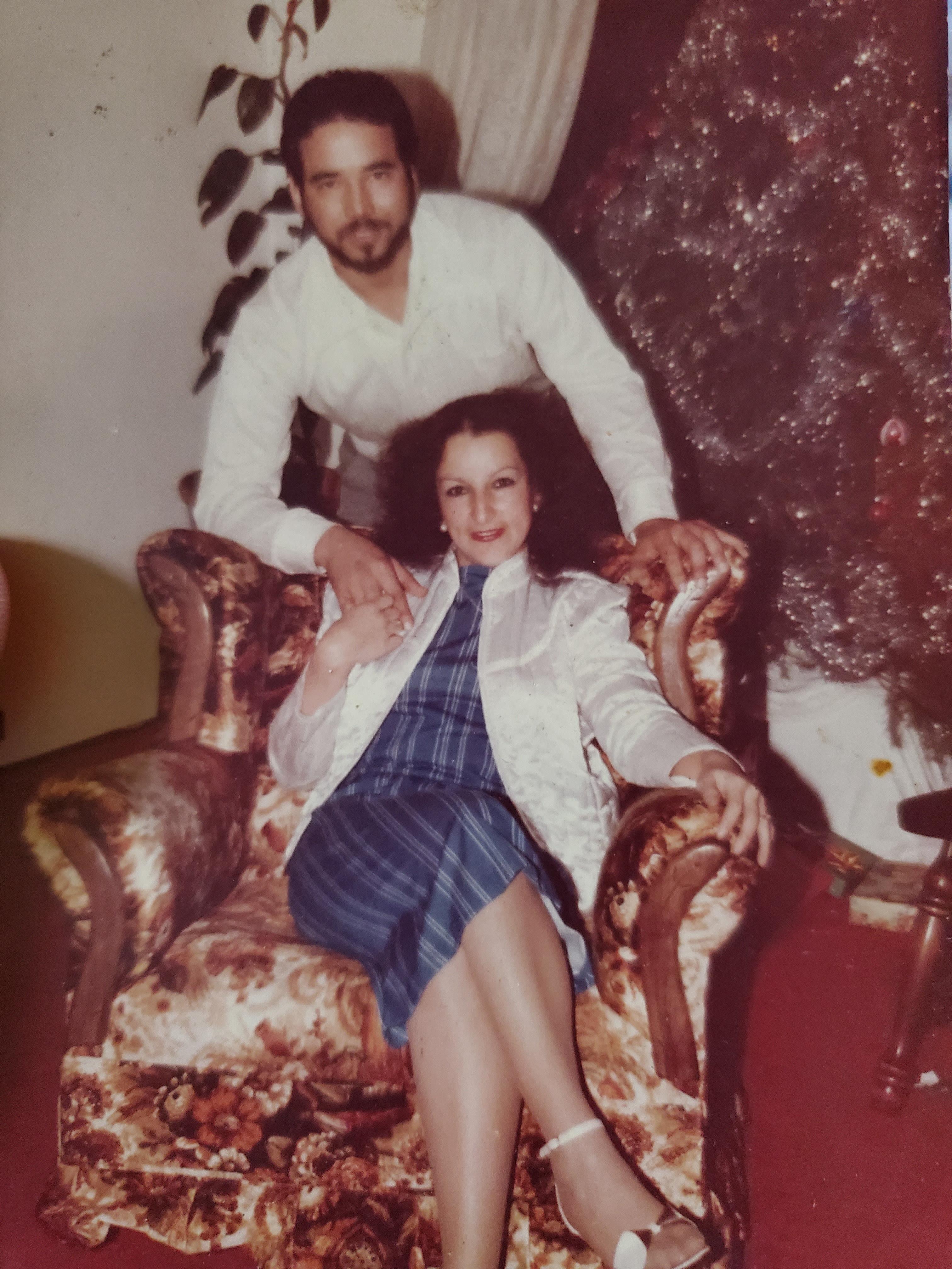 A couple poses casually on an ornate armchair, showcasing their smiles and festive attire at home.