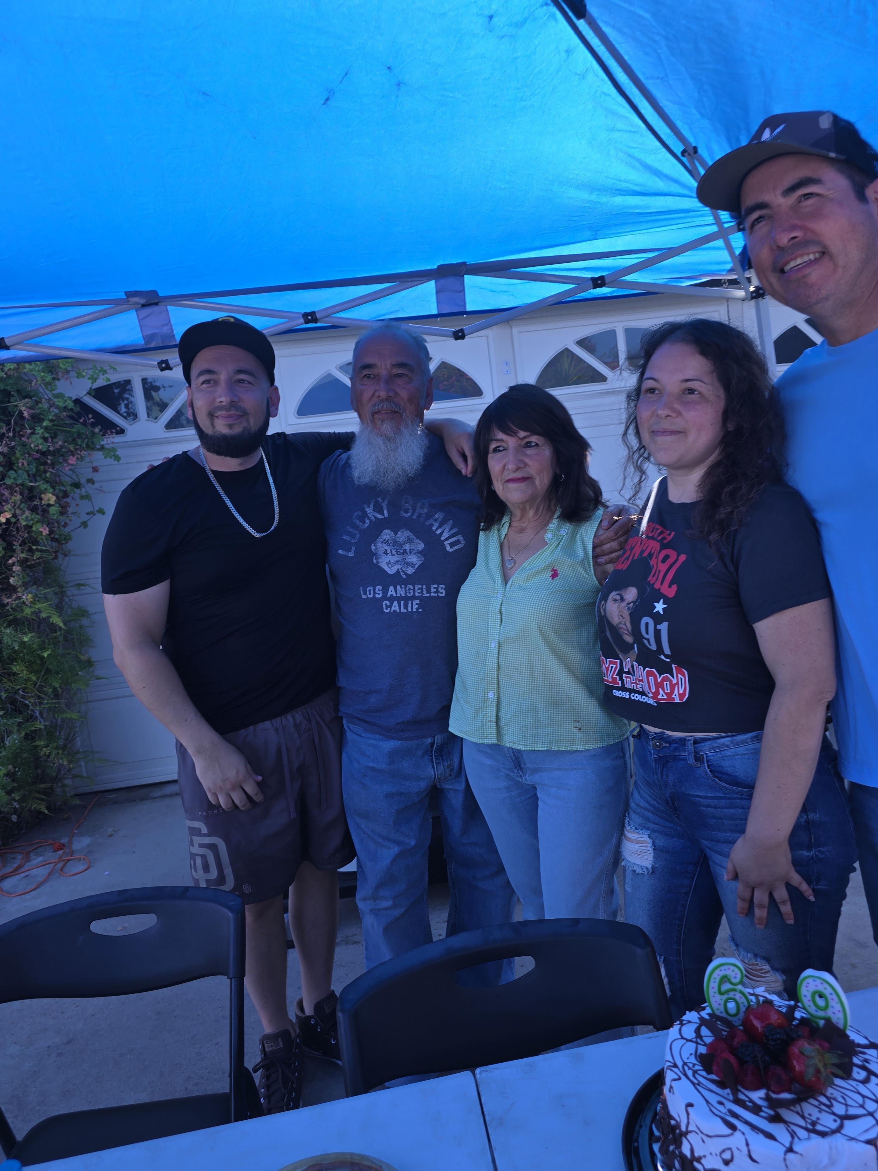Group of five people pose happily together under blue canopy at birthday celebration with cake.
