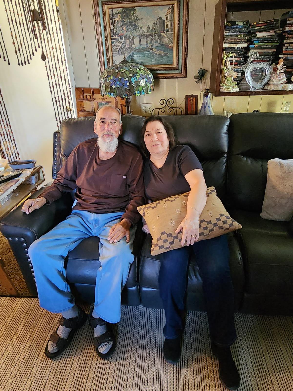An elderly couple sits on a dark sofa, sharing smiles and warmth in their cozy living room space.