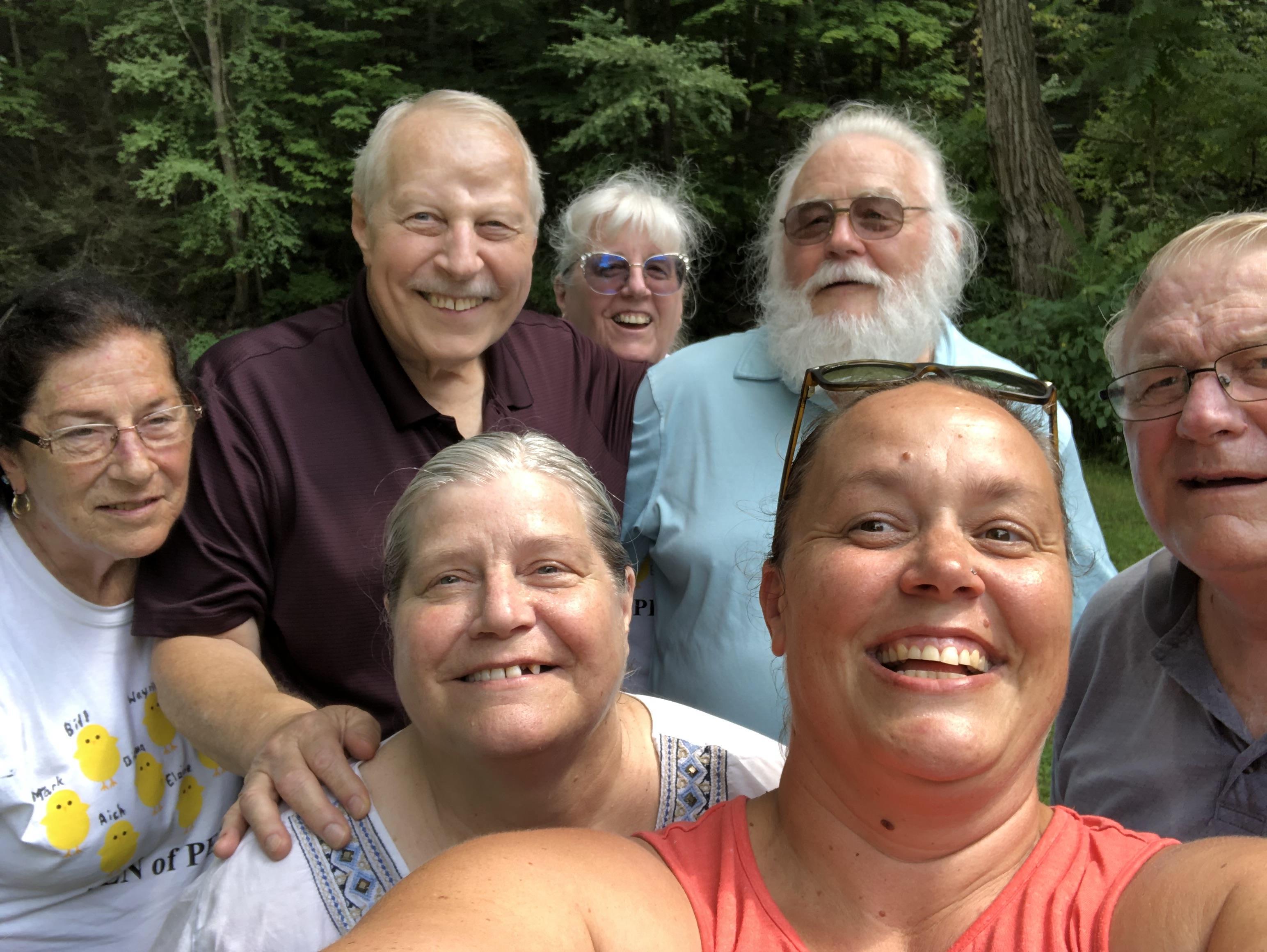 A group of friends smiles and poses for a selfie in a lush green forest during summer.