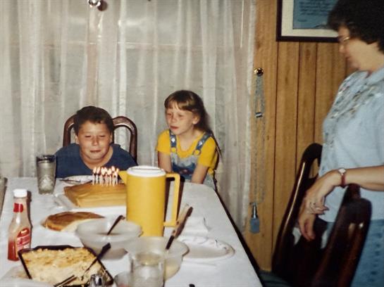 A group of kids sitting at a table with a cake and a man in the background