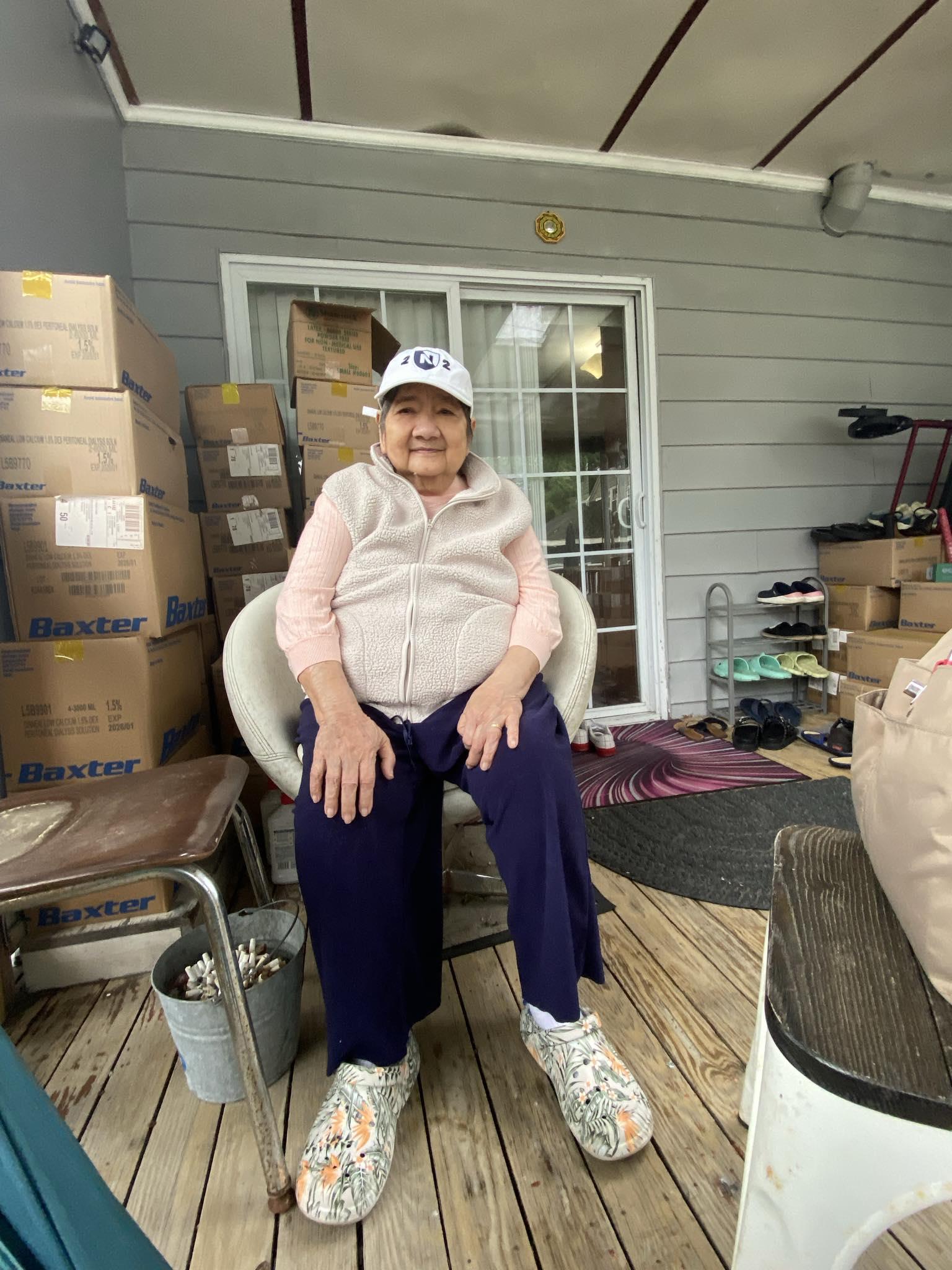 An elderly woman in casual attire relaxes on a porch amidst stacked boxes and household items.