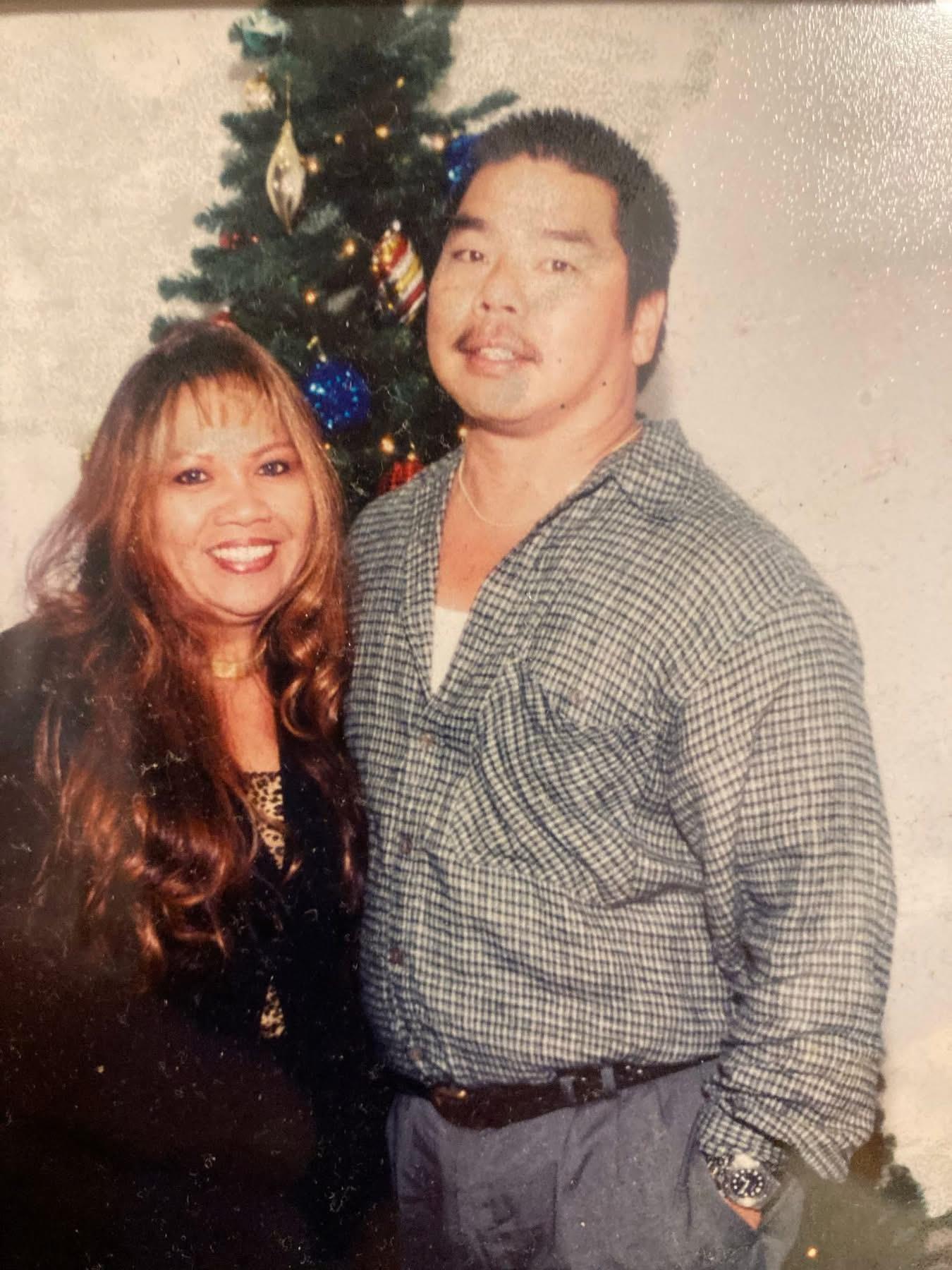 A couple poses joyfully with a decorated Christmas tree behind them during the holiday season.