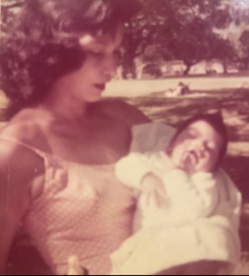 A mother lovingly holds her baby in a serene park setting under clear skies.