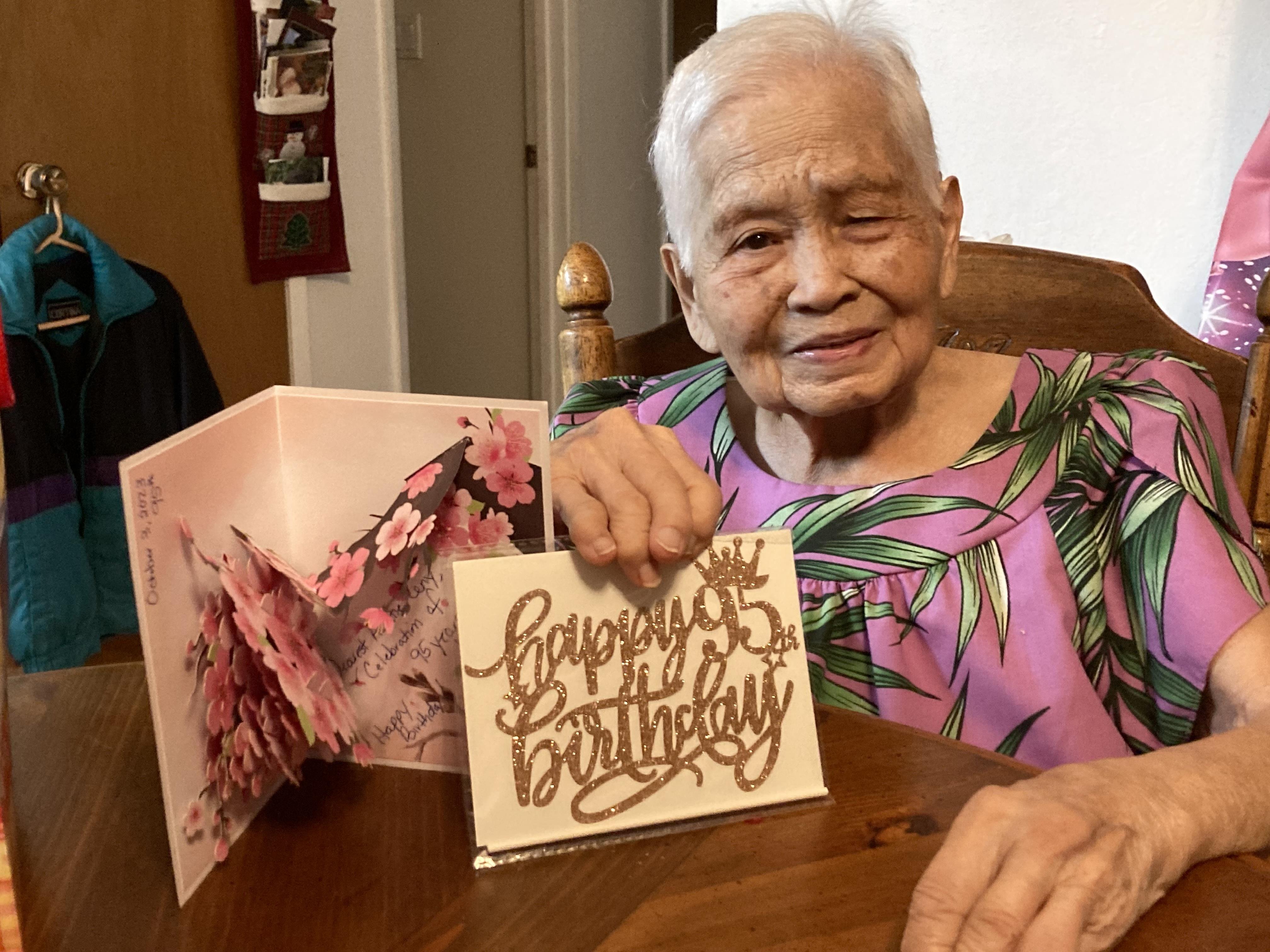 An elderly woman smiles as she holds a birthday card surrounded by home decor and warmth.