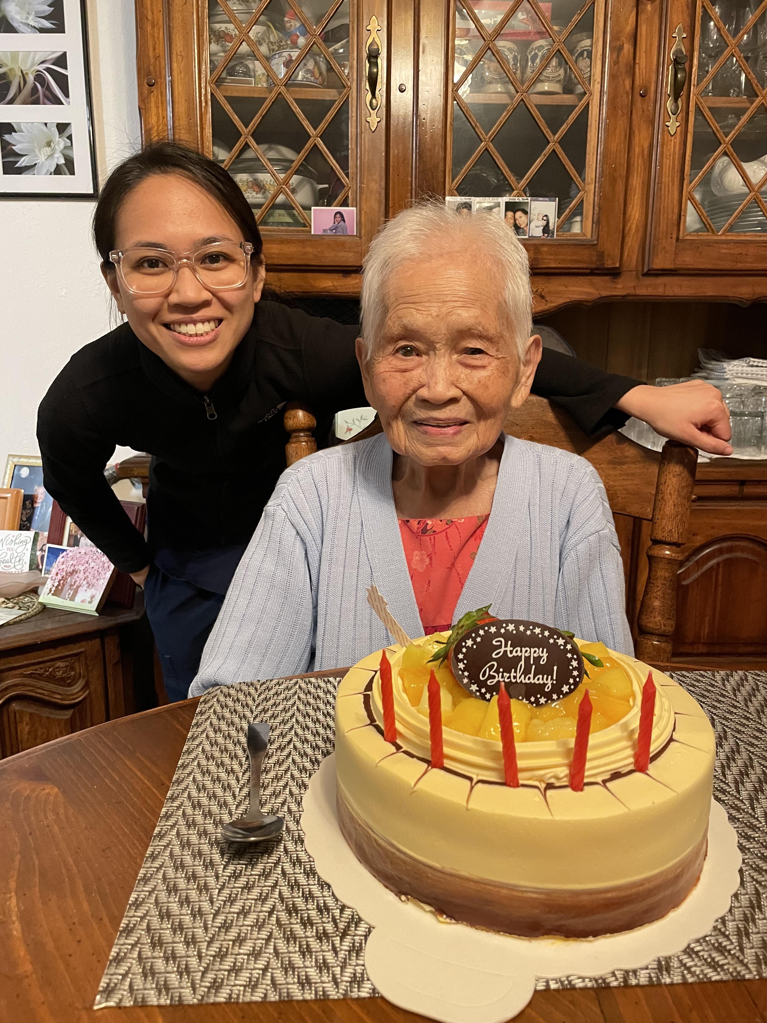 Grandmother smiles warmly at her birthday cake while granddaughter stands beside her.