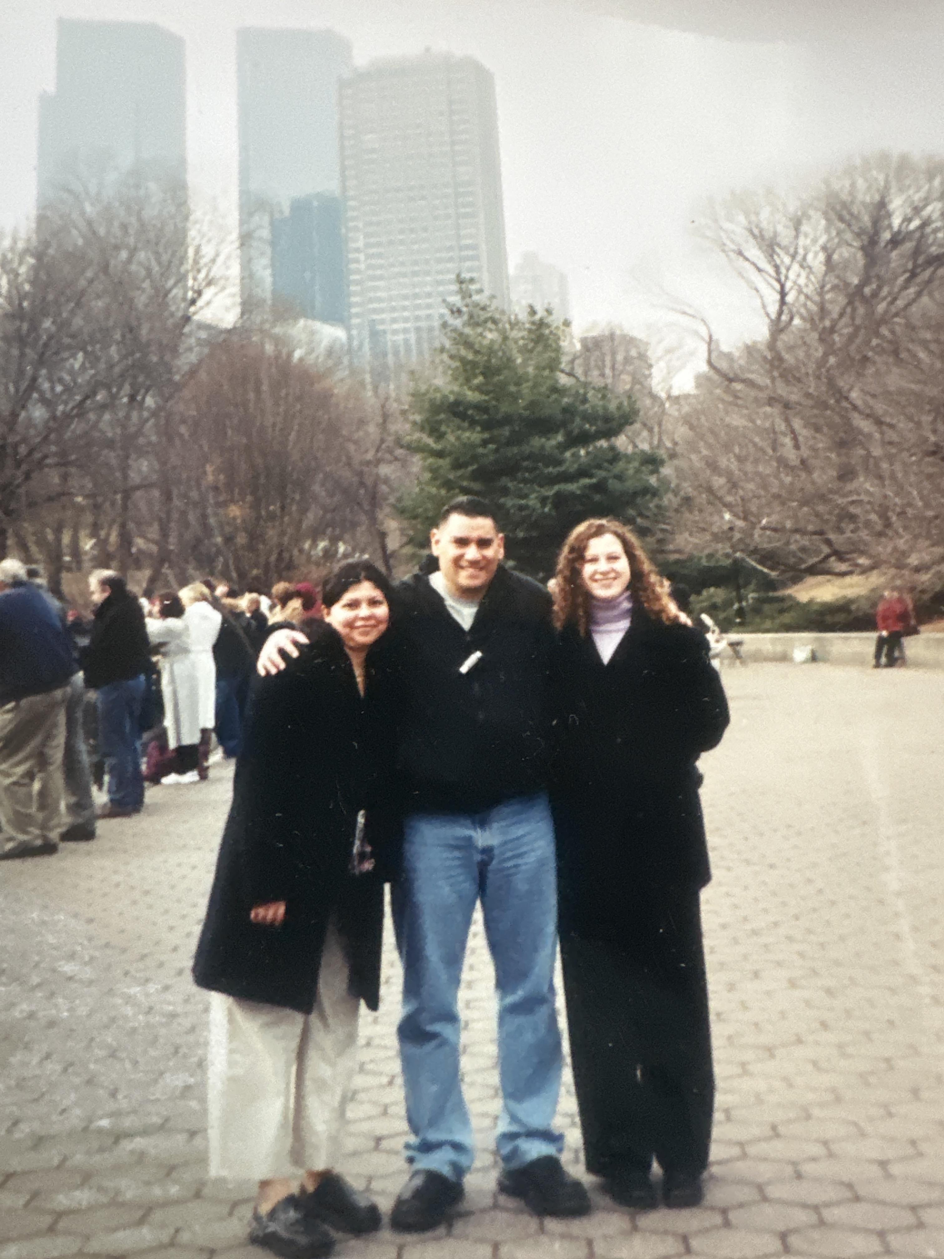 Three friends stand together in a park with a city skyline behind them on a chilly day.