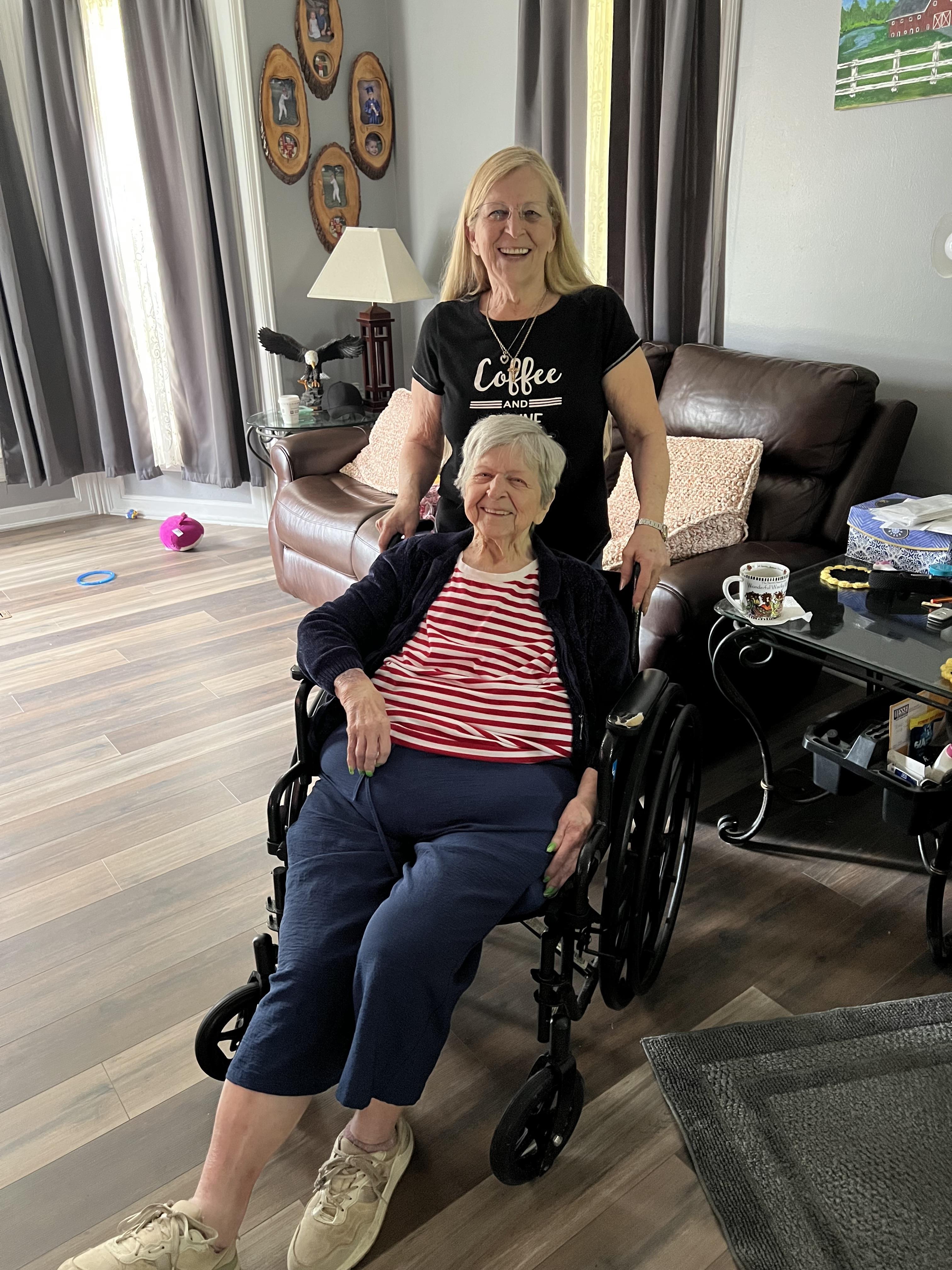 A caregiver smiles while assisting an elderly woman in a wheelchair in a warm living area.