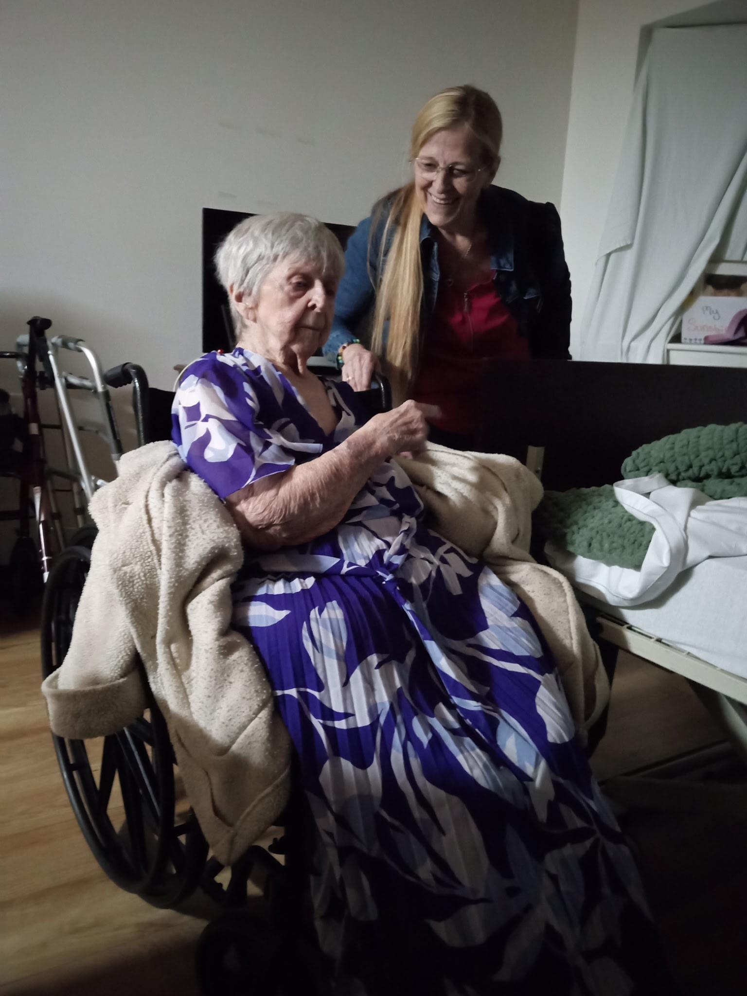 An elderly woman in a floral dress smiles as a caregiver chats with her in a warm room.