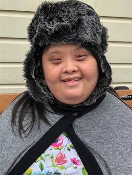 A cheerful individual poses in a furry hat and a gray cape, enjoying a relaxed moment outdoors.