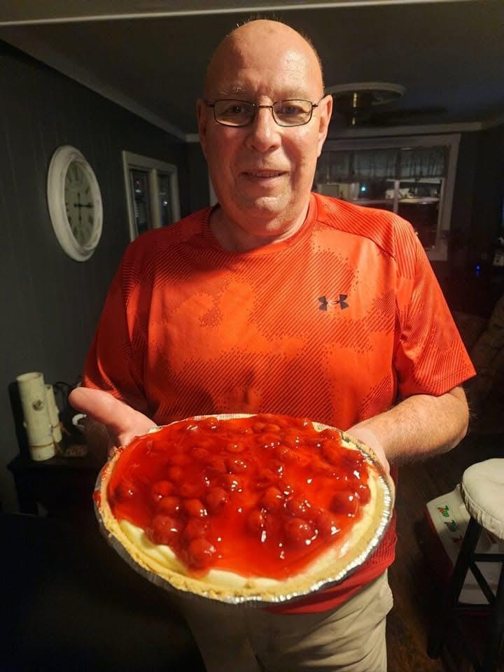A man smiles as he presents a cherry pie in a warm, inviting living room.