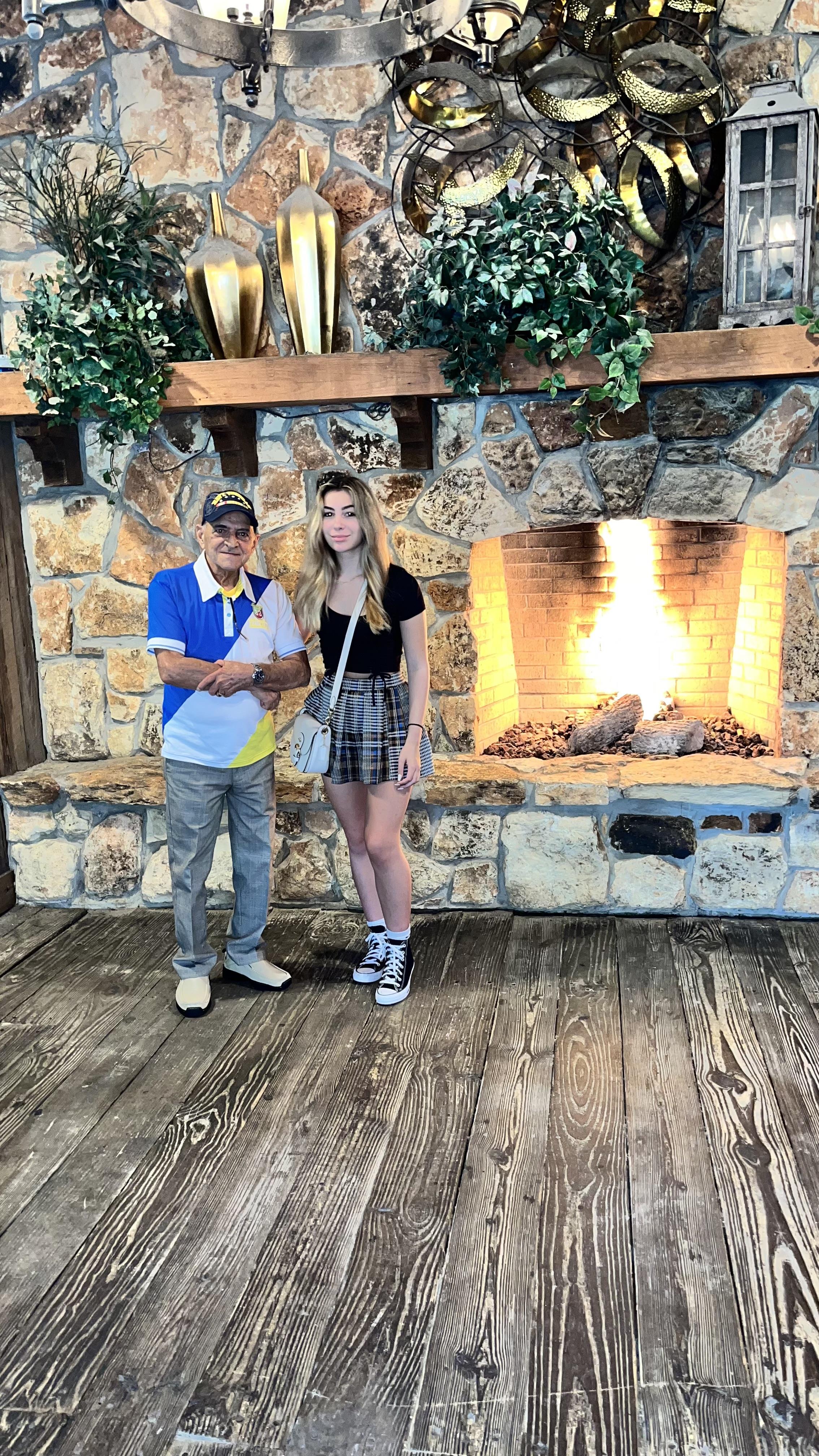 Two people pose together near a warm stone fireplace in a rustic lodge setting.