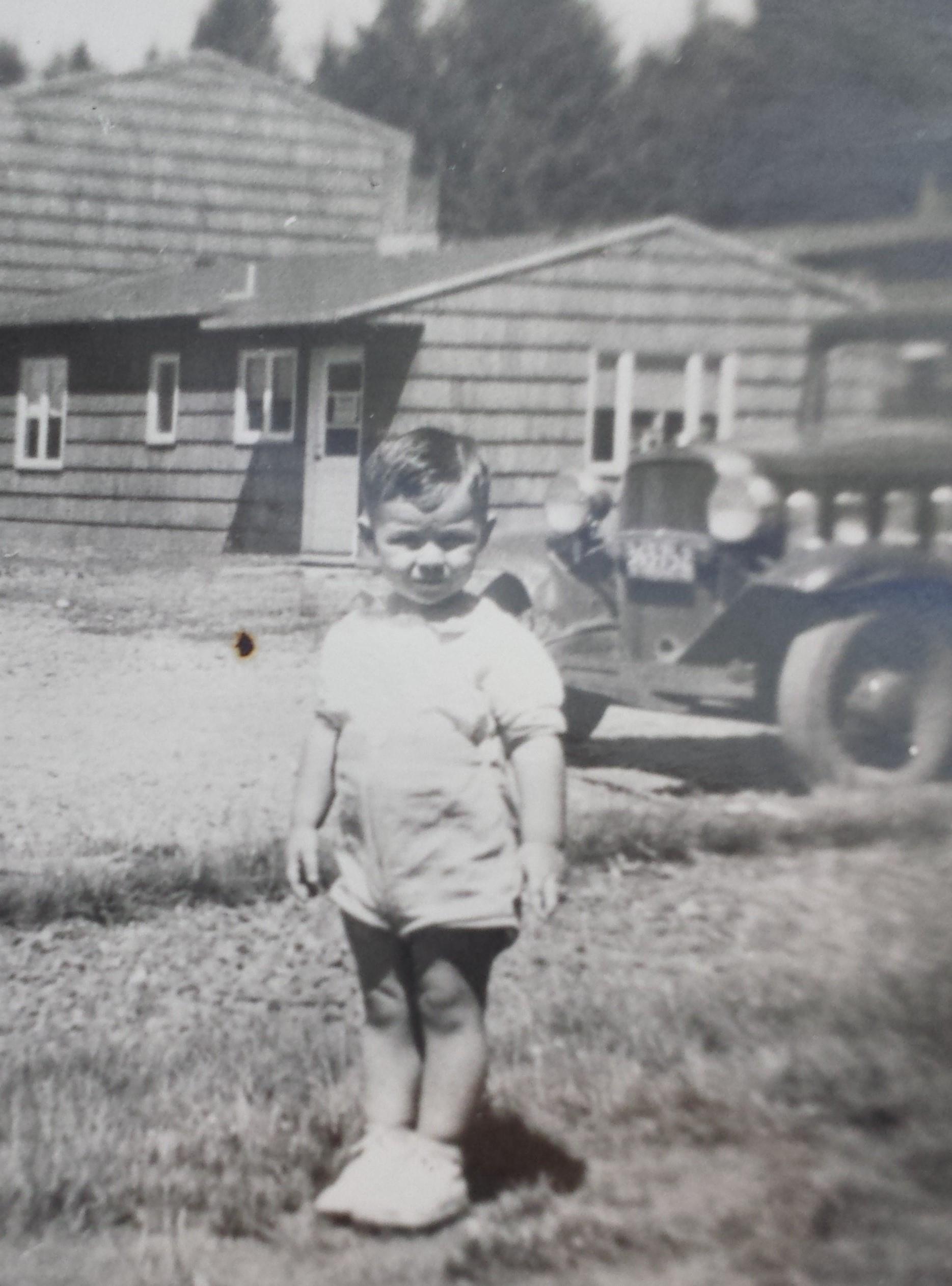 Young child poses outside a rustic house with a classic car nearby during sunny weather.