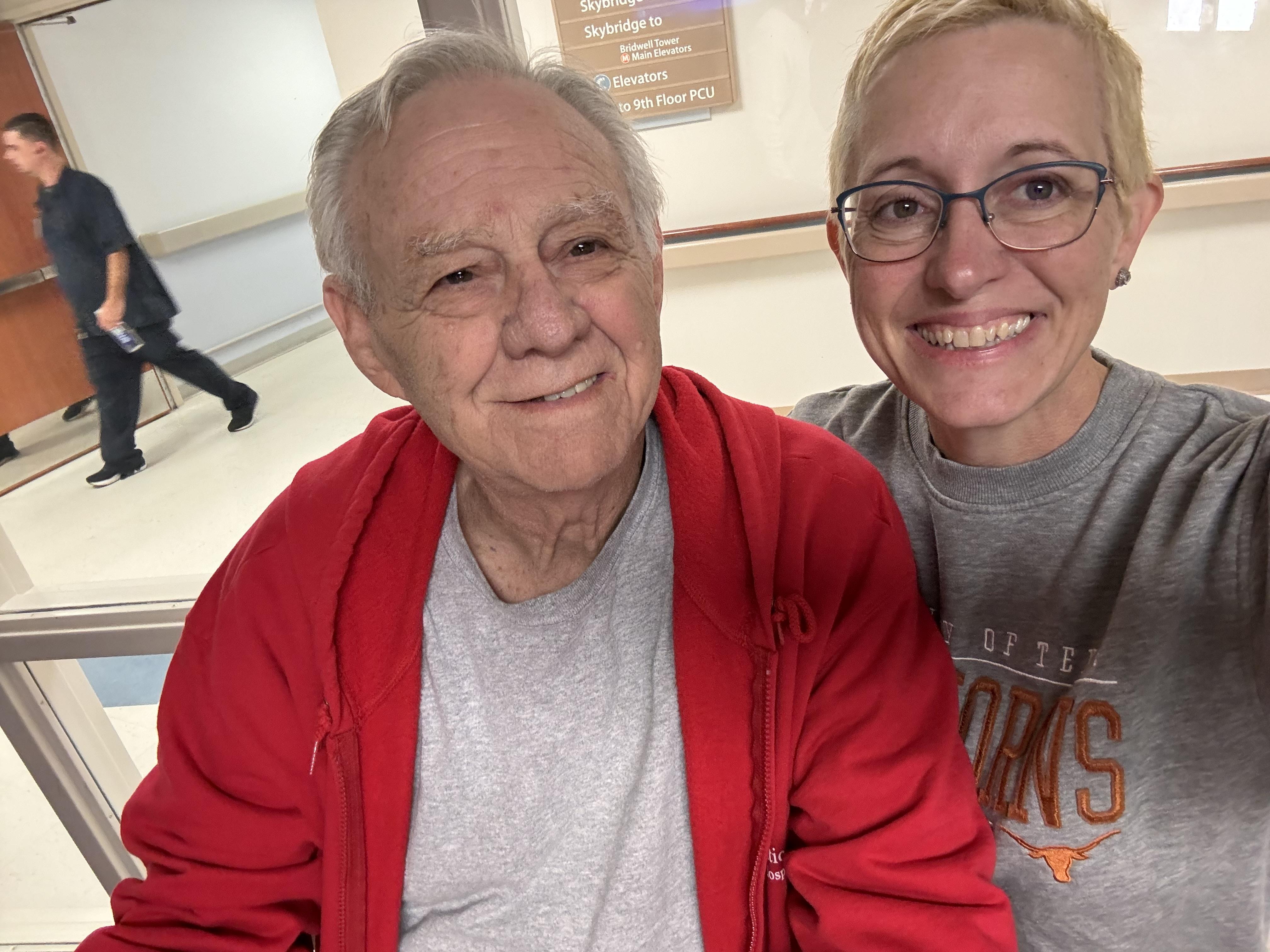 A caregiver takes a joyful selfie with an elderly man in a hospital corridor during a visit.