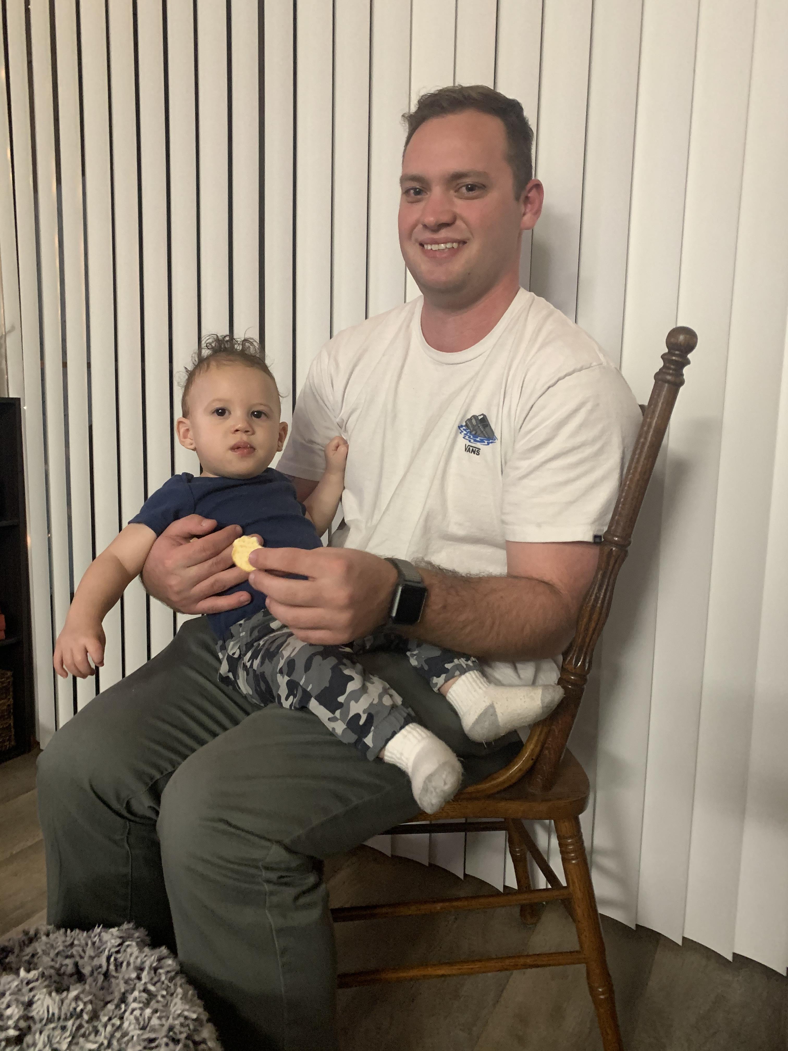 A father sits in a chair with his baby enjoying a snack in a cozy room.