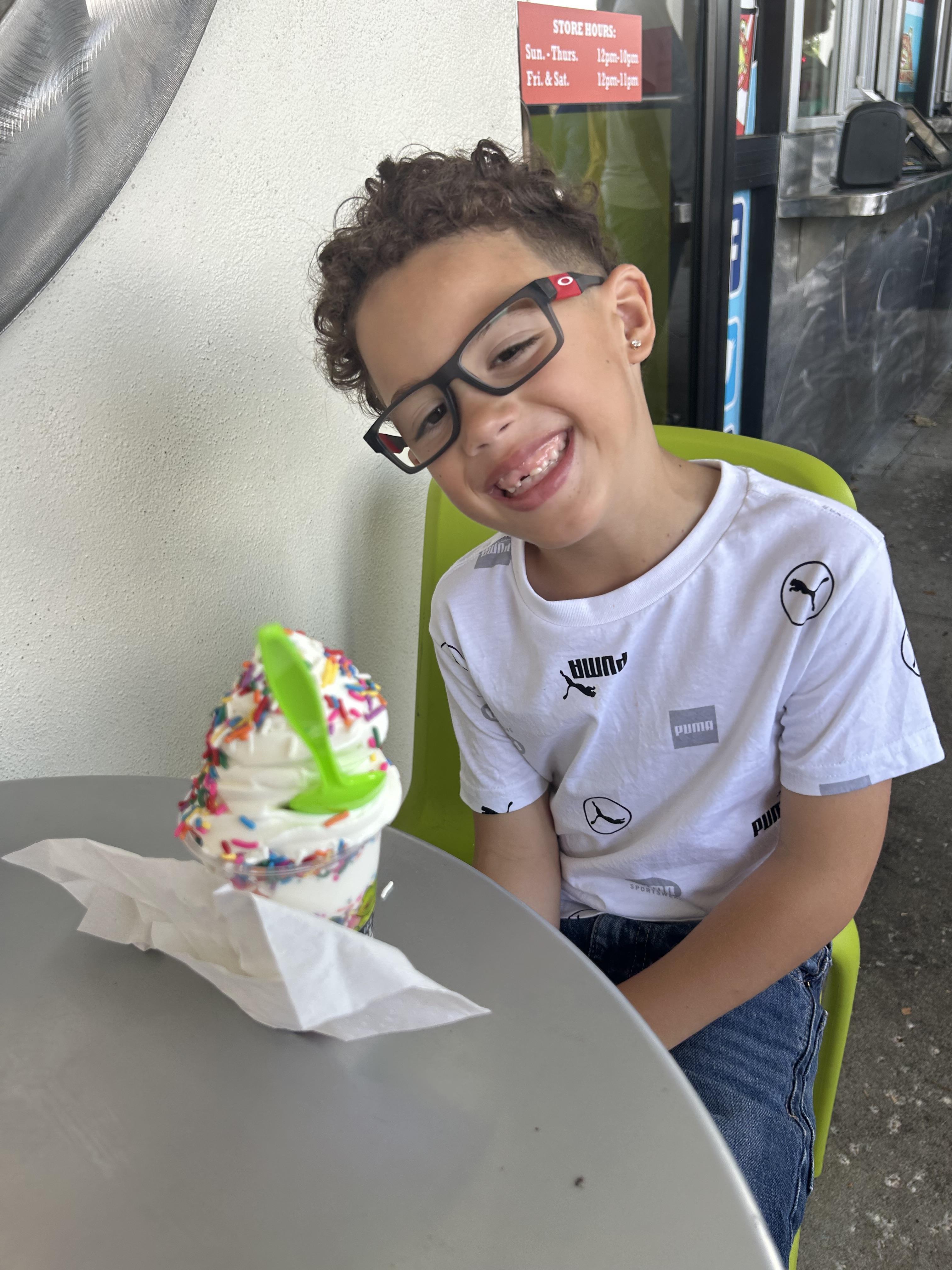A young boy smiles at a table, enjoying a sprinkles-topped ice cream.