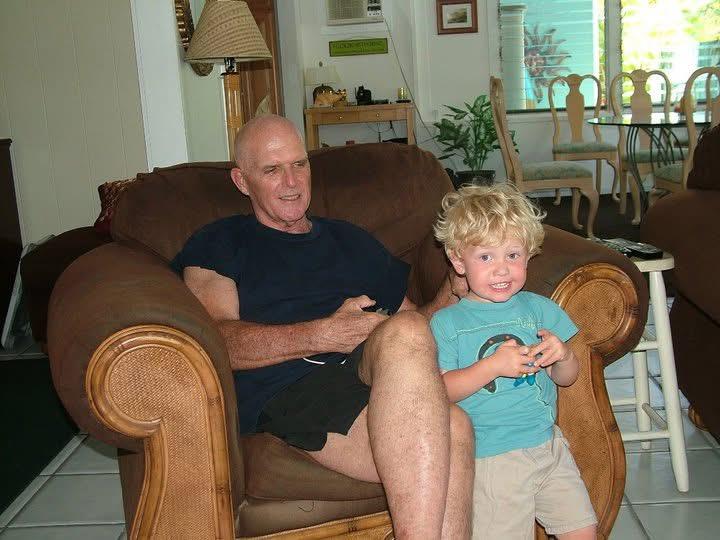 An elder man and young boy sit together on a couch, both engaged in playing video games with joy.