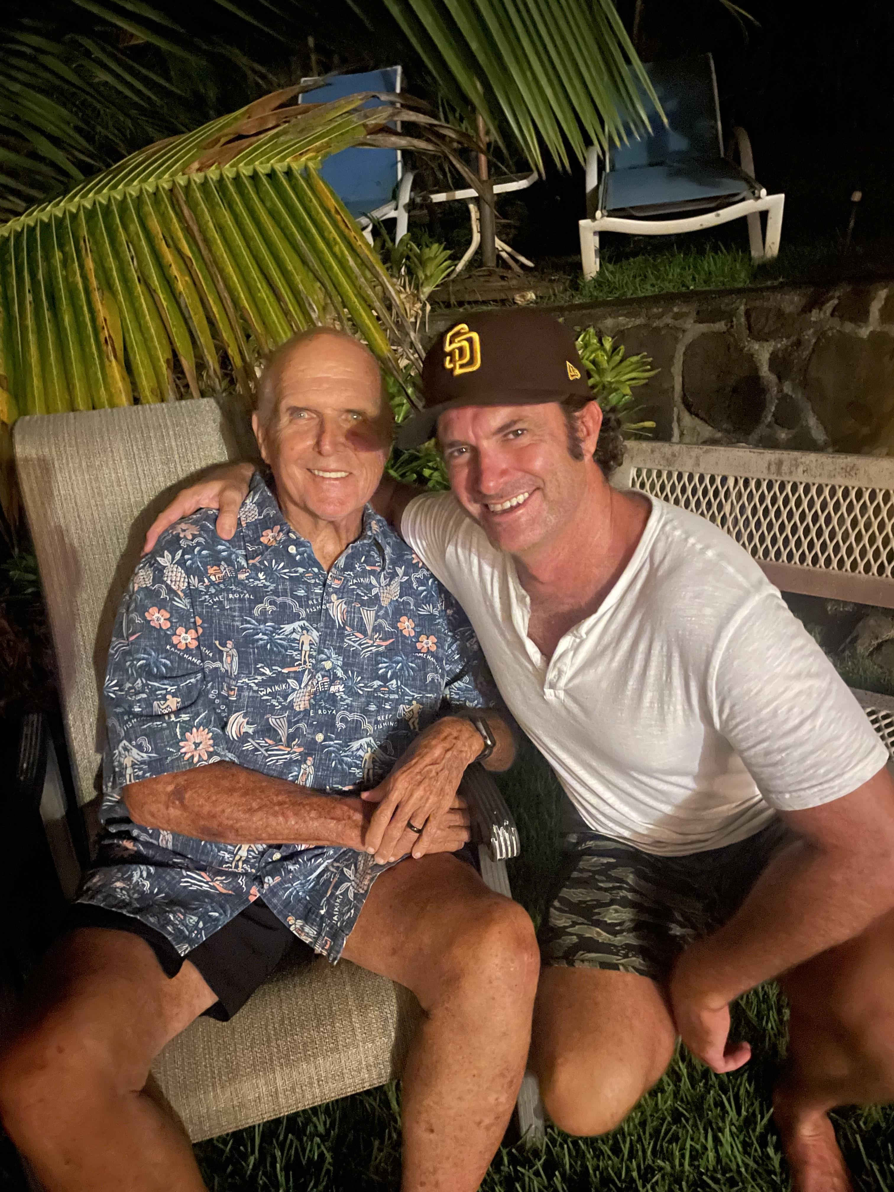 Two men smile warmly while sitting outdoors, enjoying each other's company under palm trees.