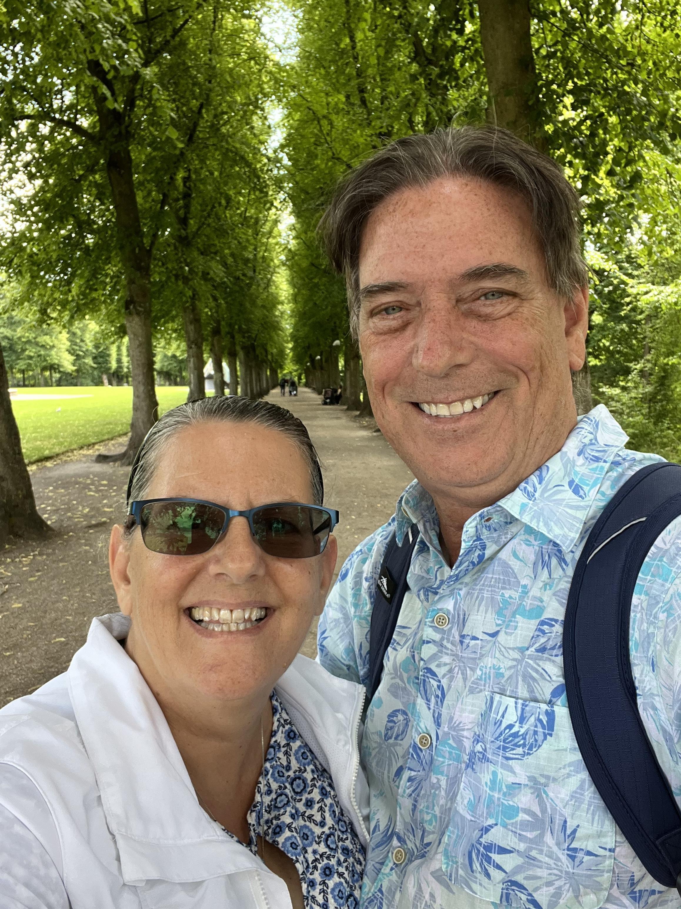 A couple smiles together while walking under tall trees in a lively park on a sunny day.