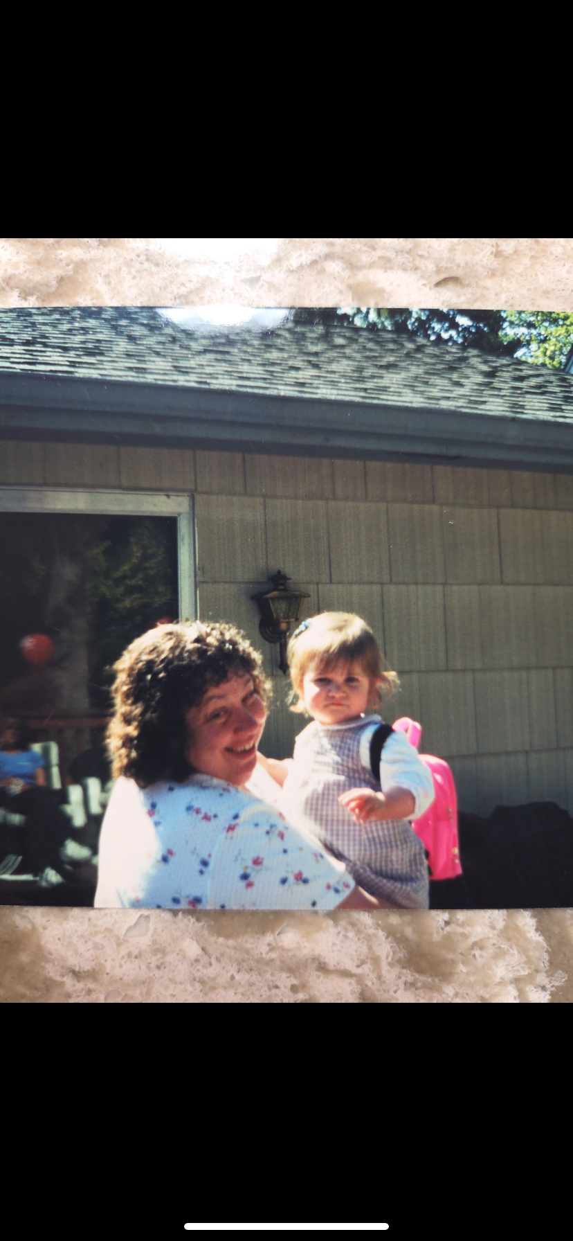 A cheerful woman embraces a toddler dressed in a striped shirt and pink backpack outside a home.