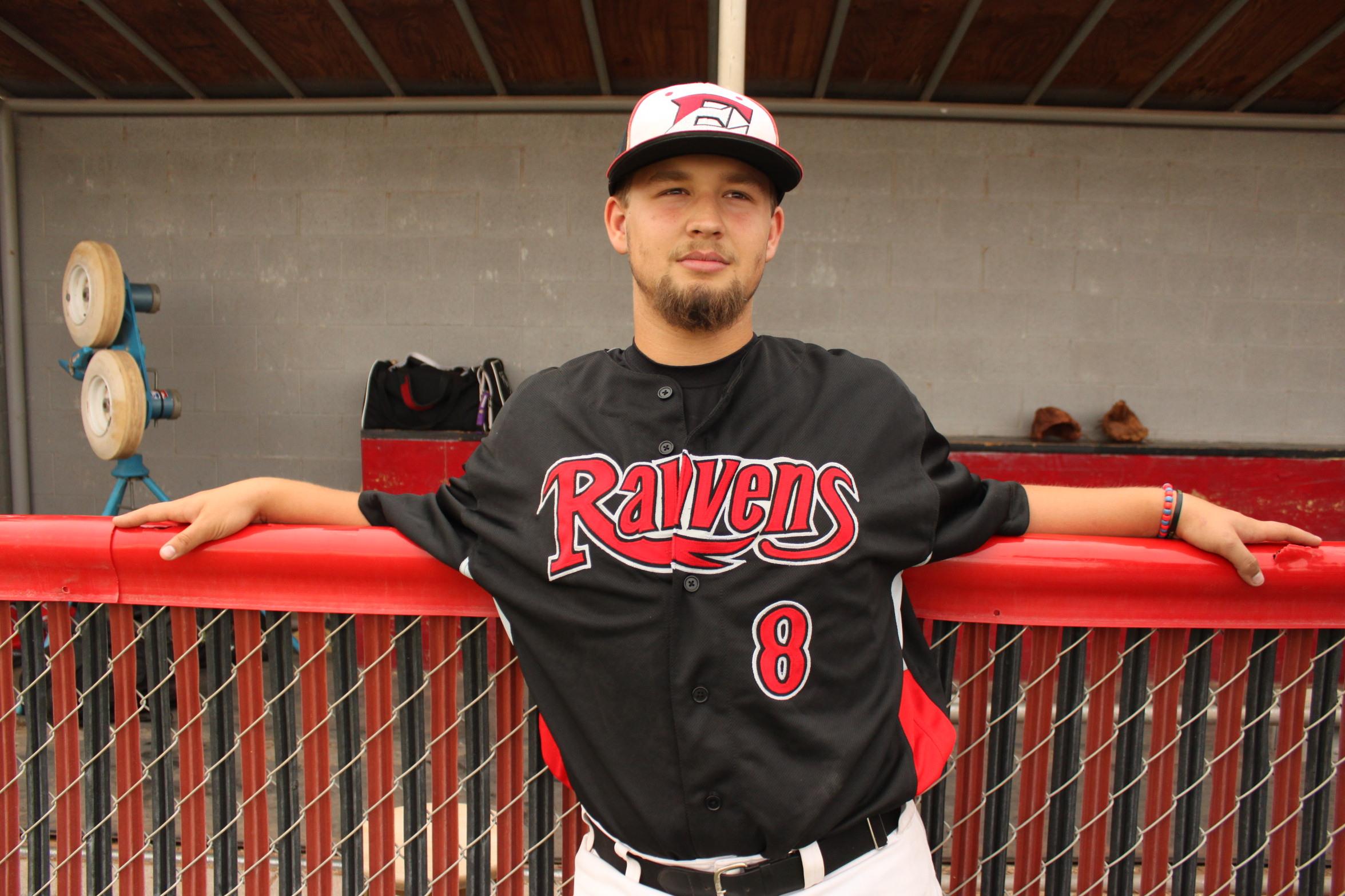 A baseball player in a black and red jersey leans on the dugout rail, smiling.