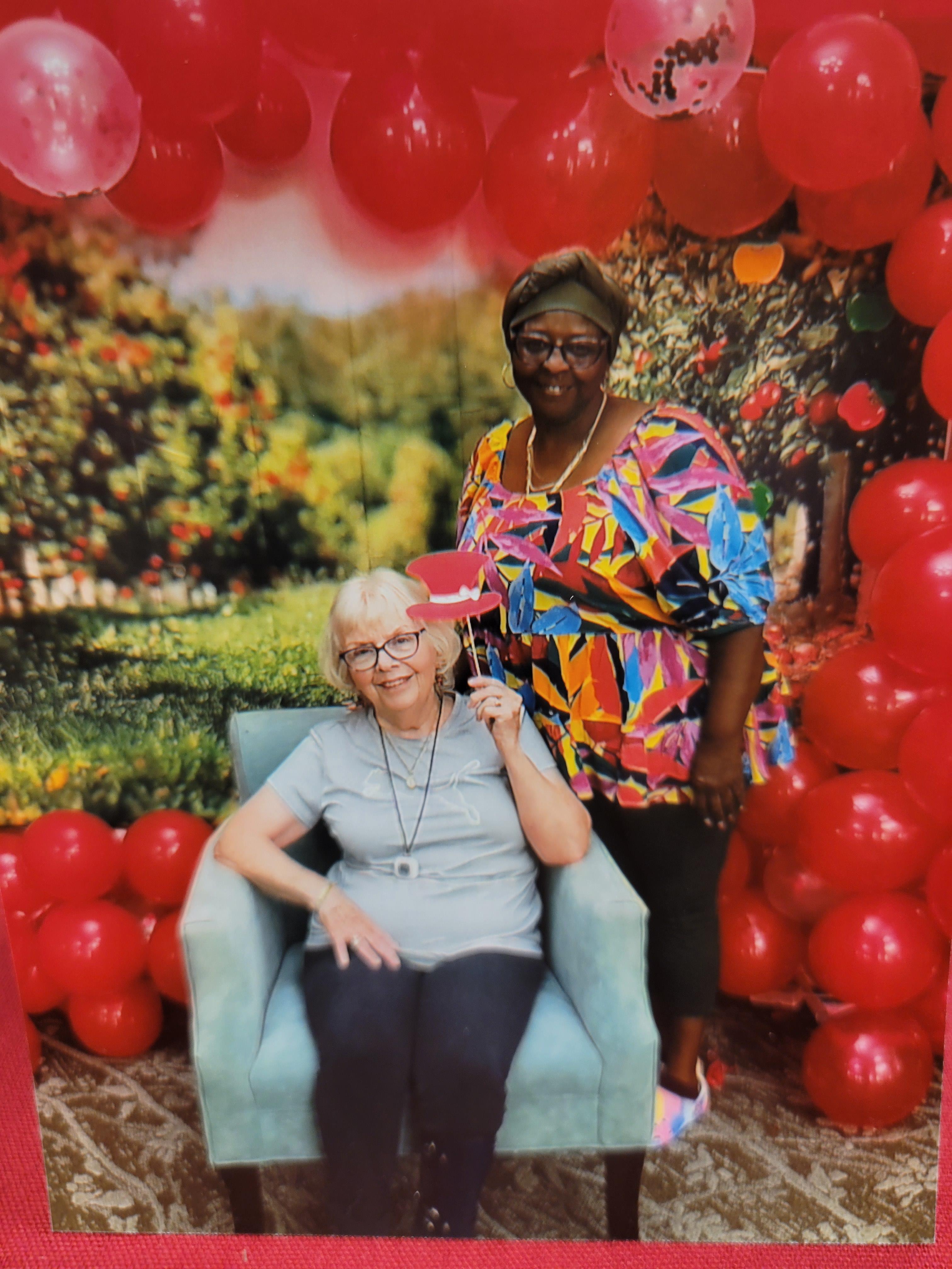 Two women pose joyfully in front of a colorful backdrop adorned with balloons and greenery.