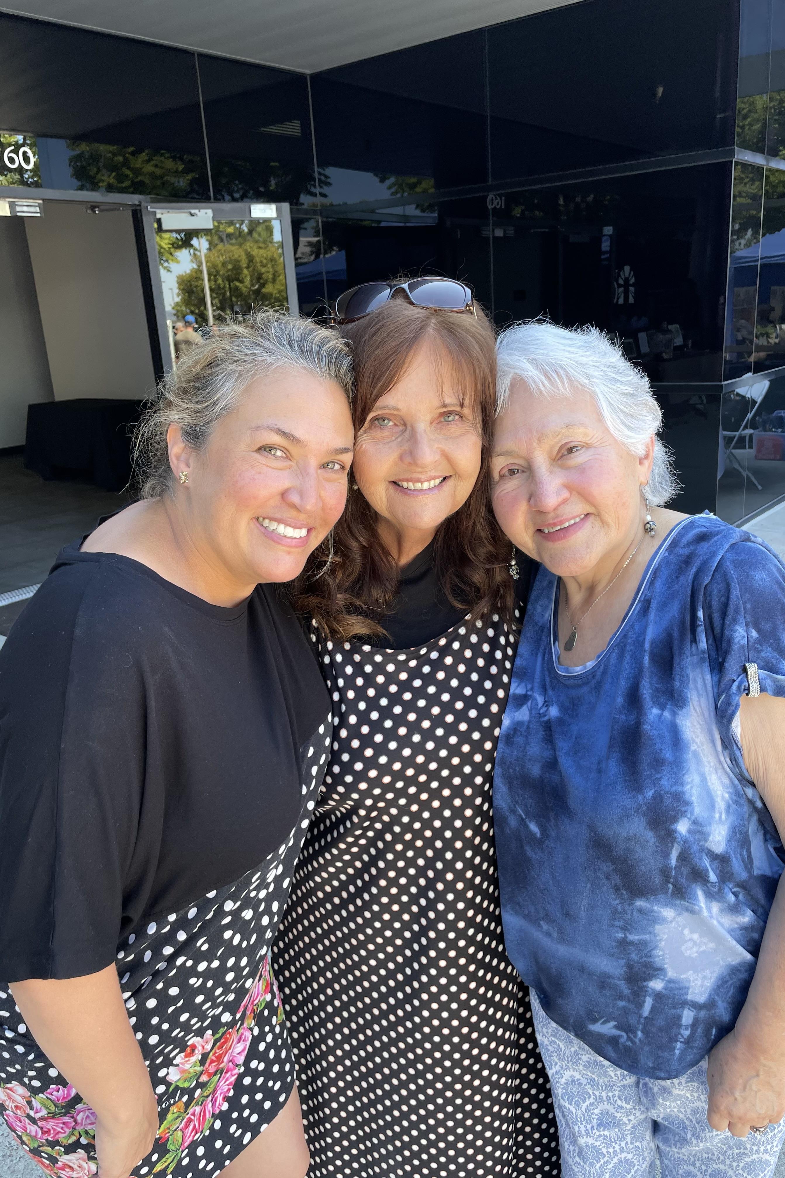 Three women smile joyfully together outside a venue during a family celebration, radiating warmth.