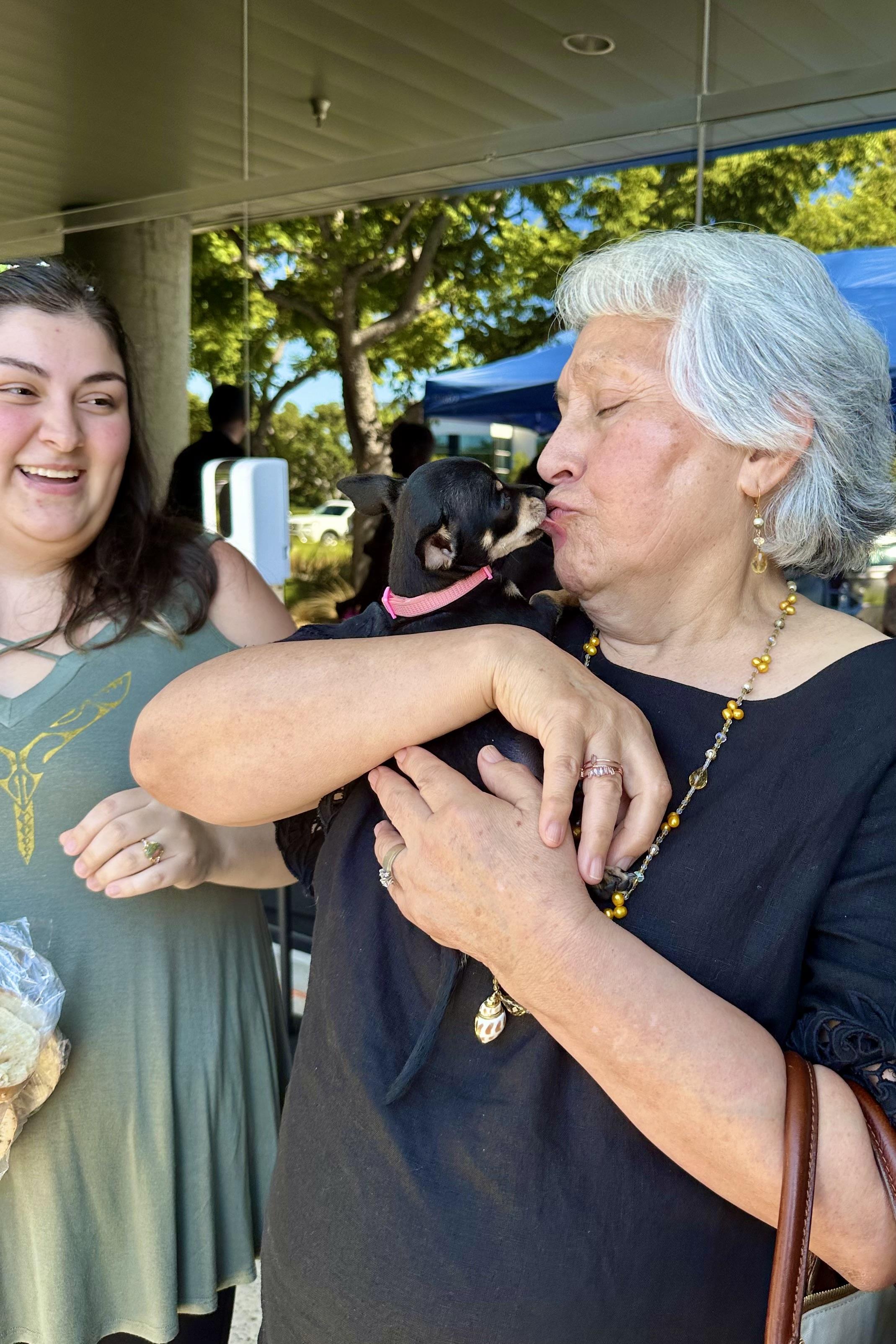 A woman cuddles a small dog as another woman shares a joyful expression nearby at an event.