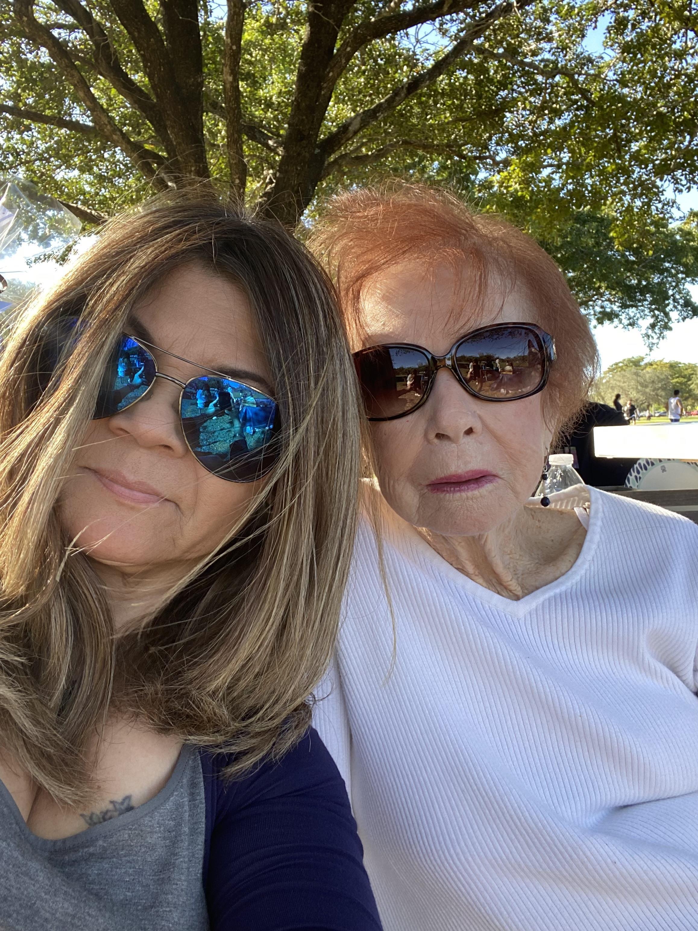 Two women sit closely together under a tree, enjoying a bright day and each other's company.