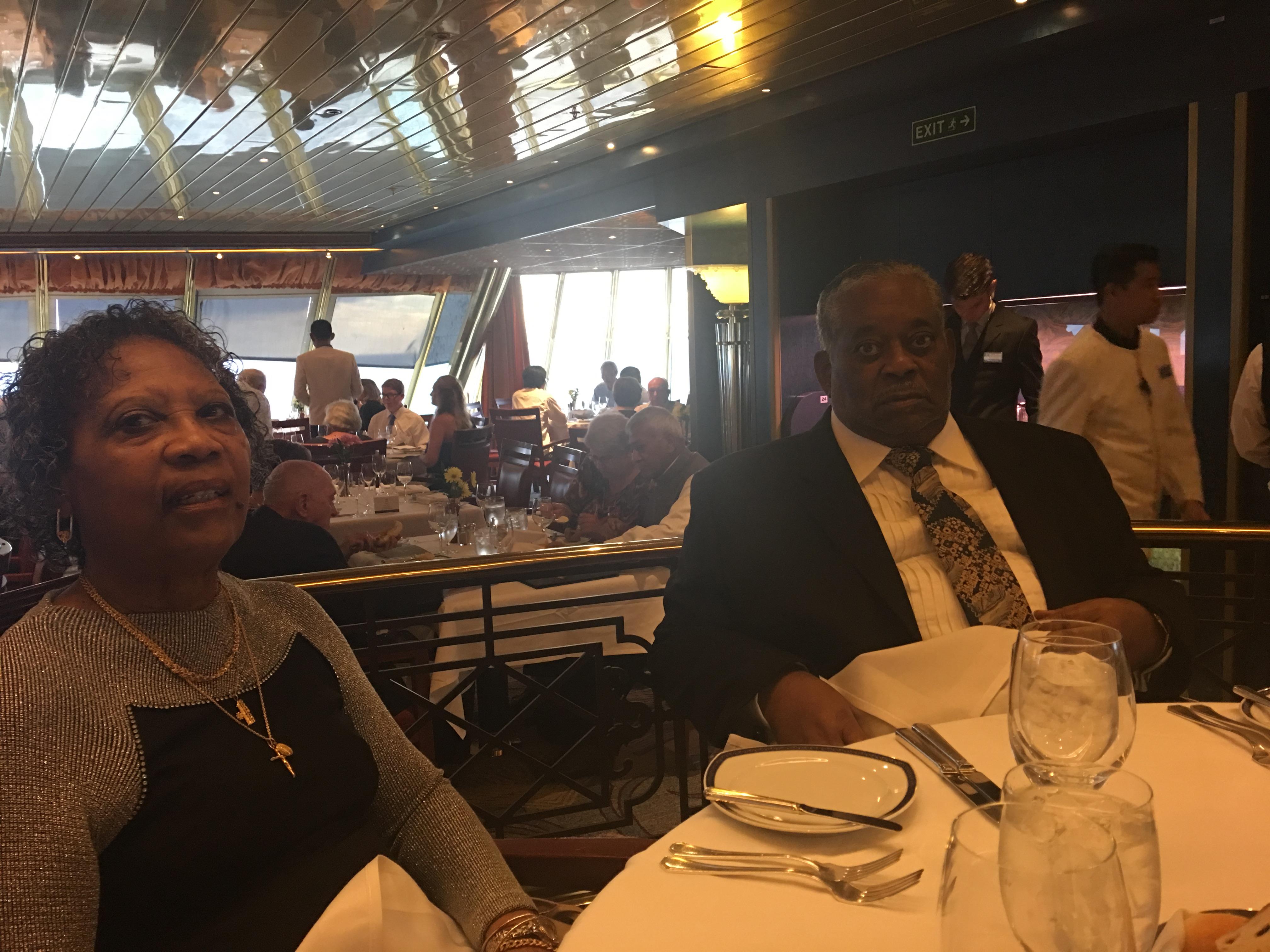 Two guests sit at a table in a cruise ship dining area, dressed for an elegant meal.