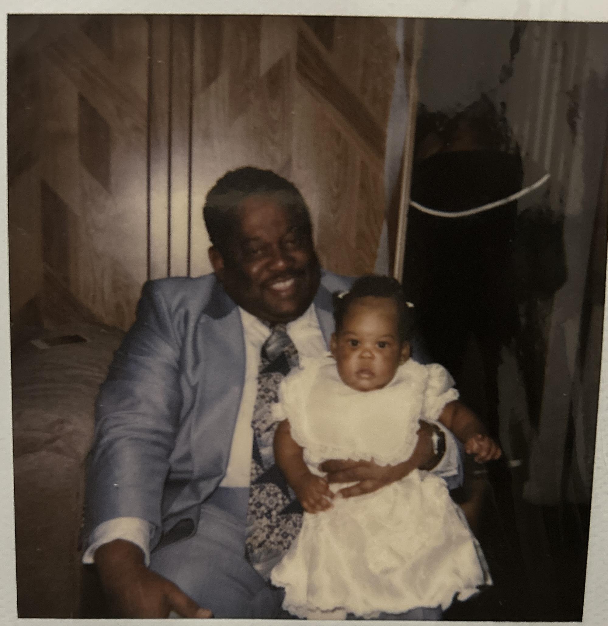 An elderly man smiles broadly while holding a baby girl wearing a white dress in a warm room.