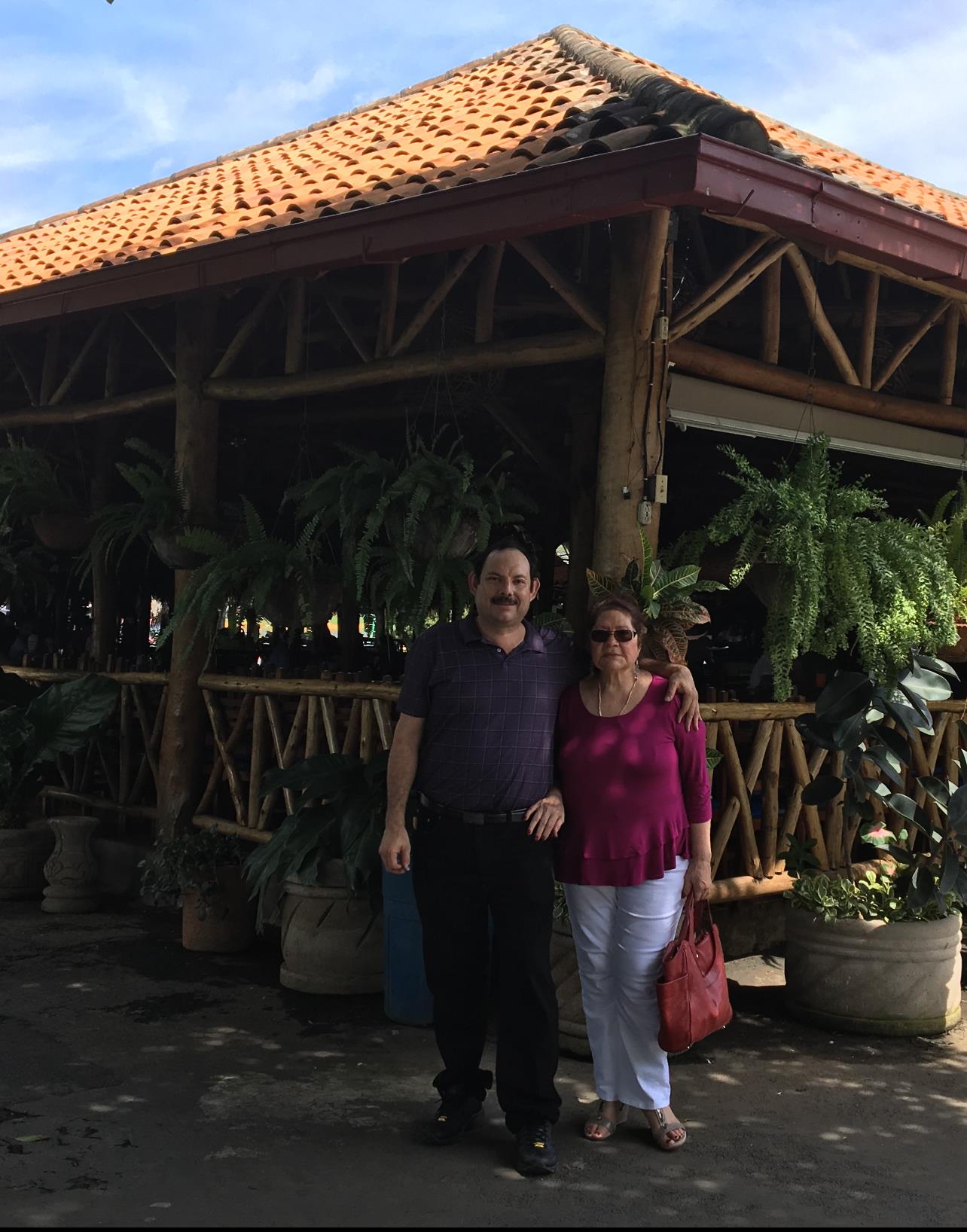 Two people stand together, smiling in front of a rustic restaurant adorned with plants.