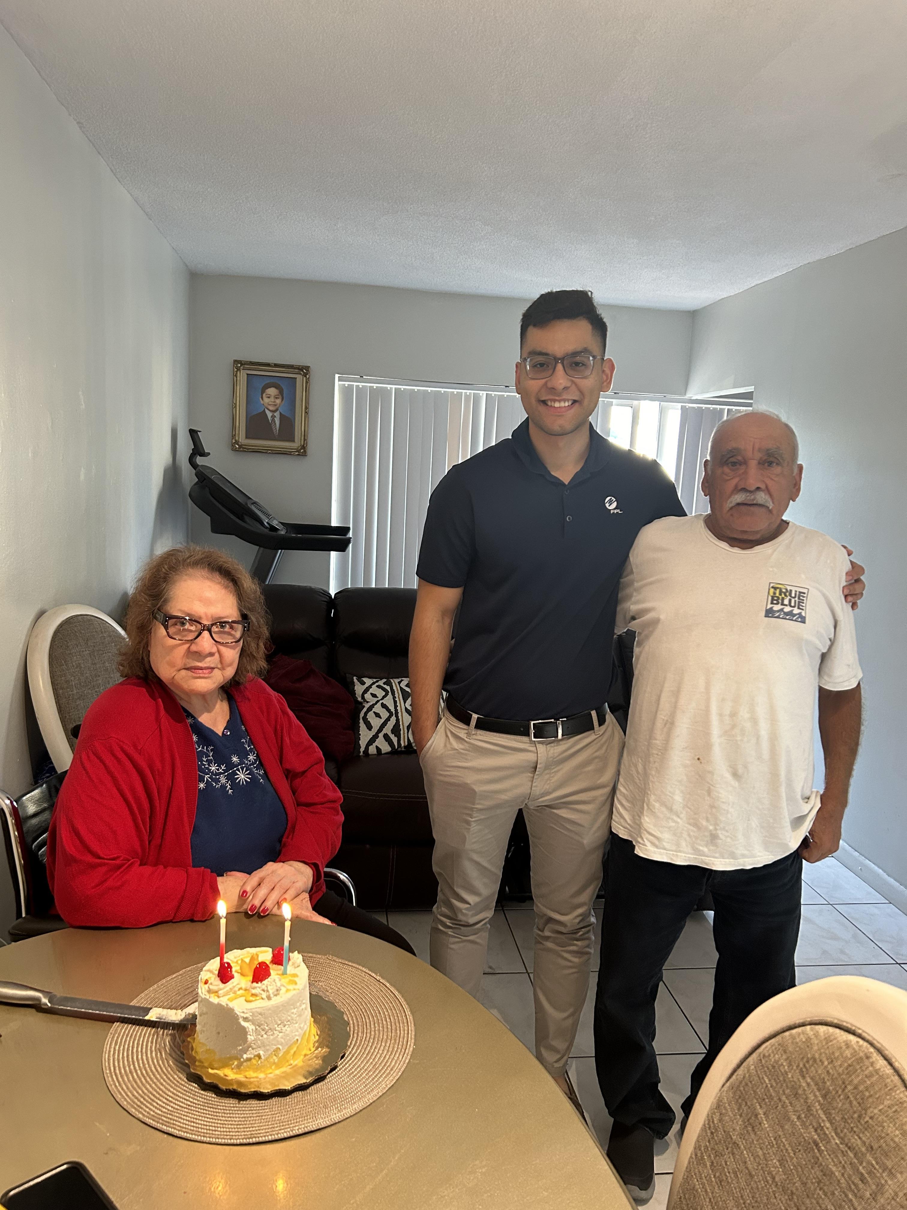 Three family members gather around a birthday cake, smiling together in a comfortable living space.