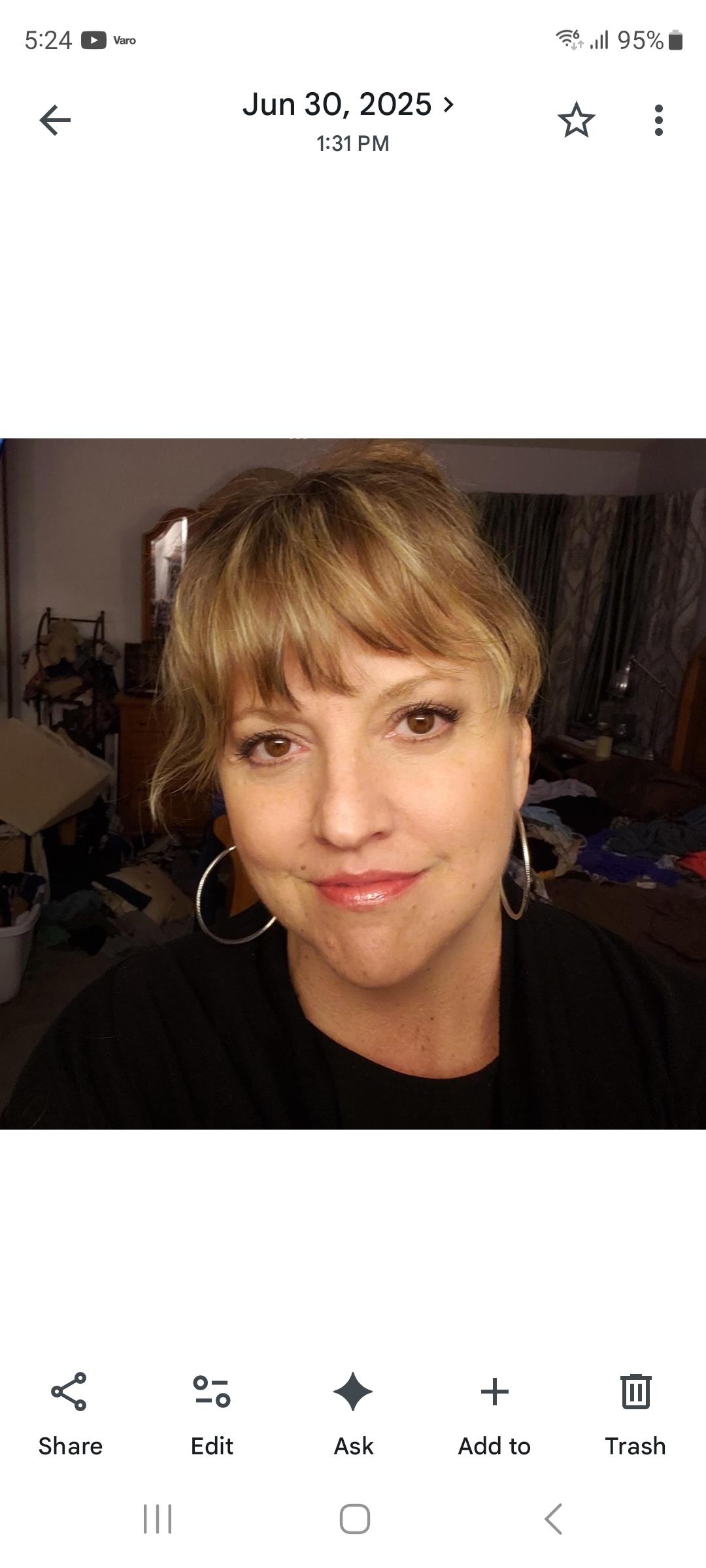 A woman with light brown hair looks directly at the camera while standing in a cluttered room.