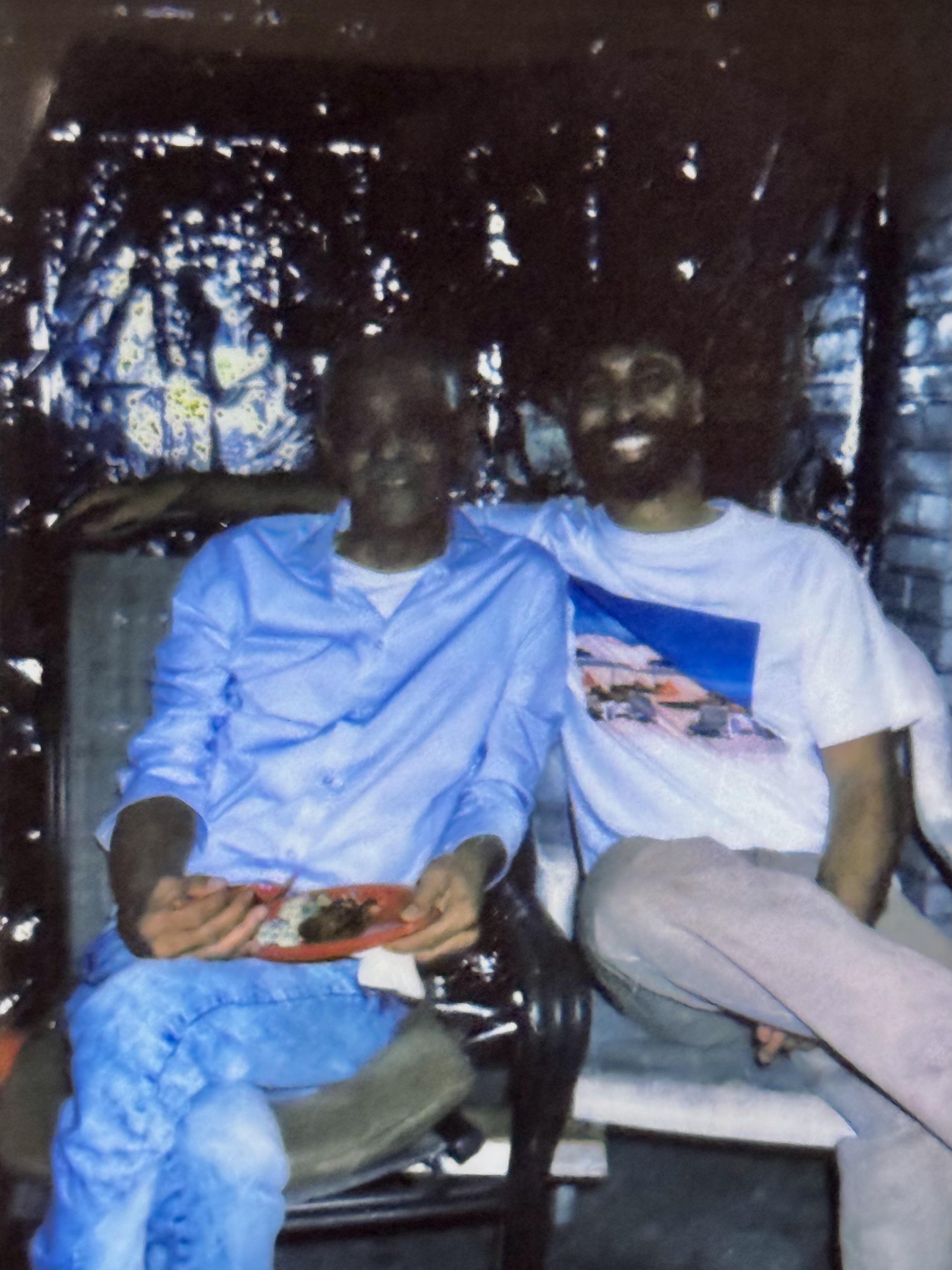Two men sit comfortably, sharing a plate of food in an intimate indoor space, laughing together.