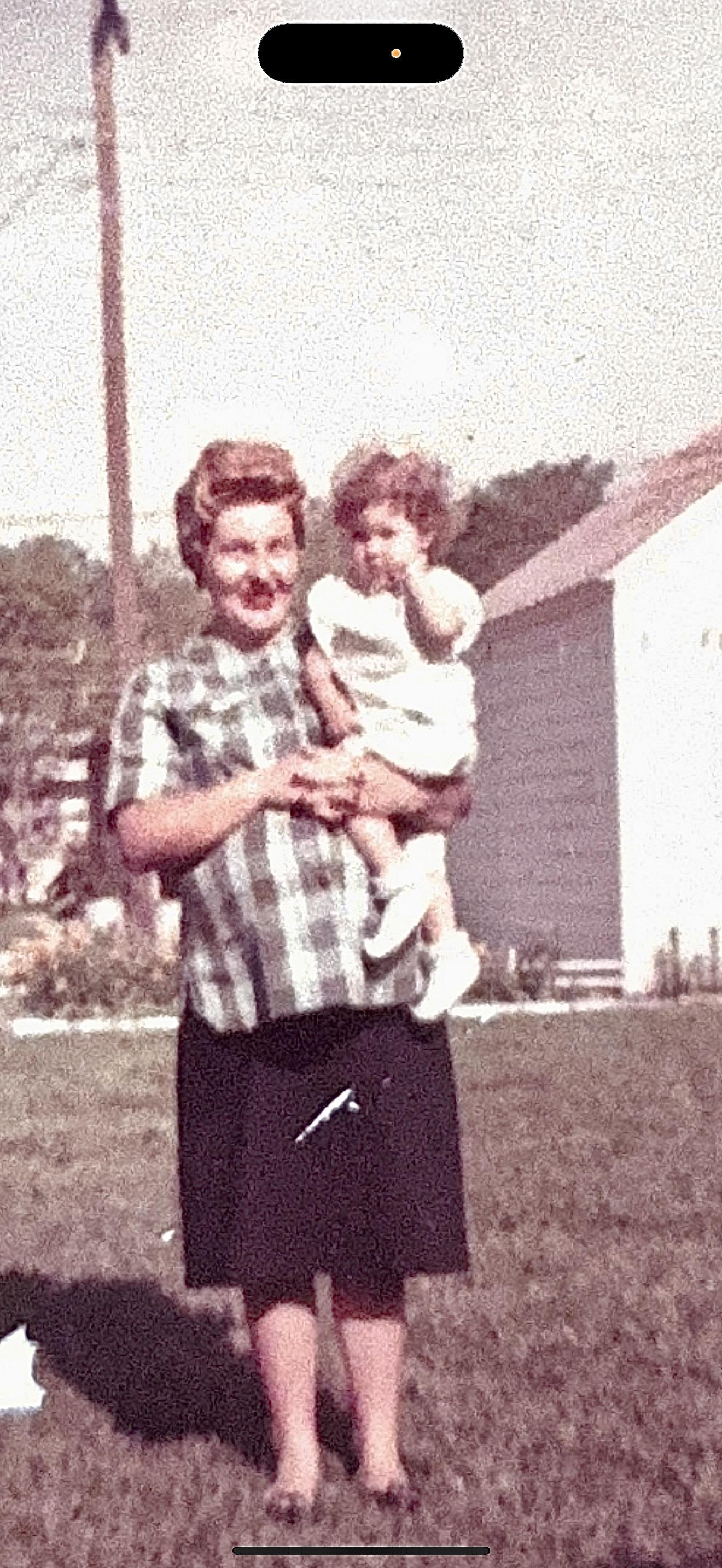 A woman proudly holds a young child in her arms, both smiling joyfully in the backyard.