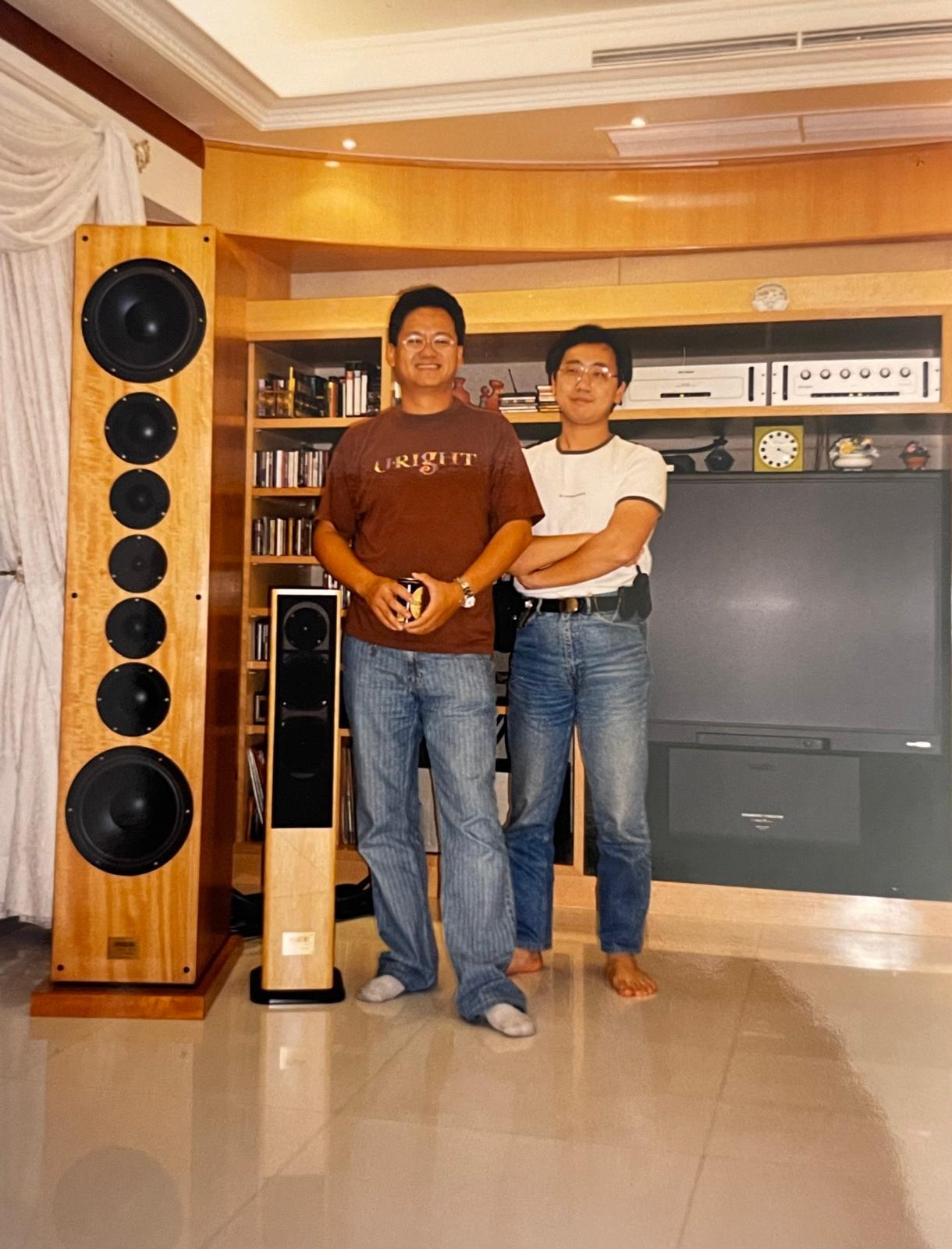 Two friends stand in a warm living room filled with audio equipment and a large television.