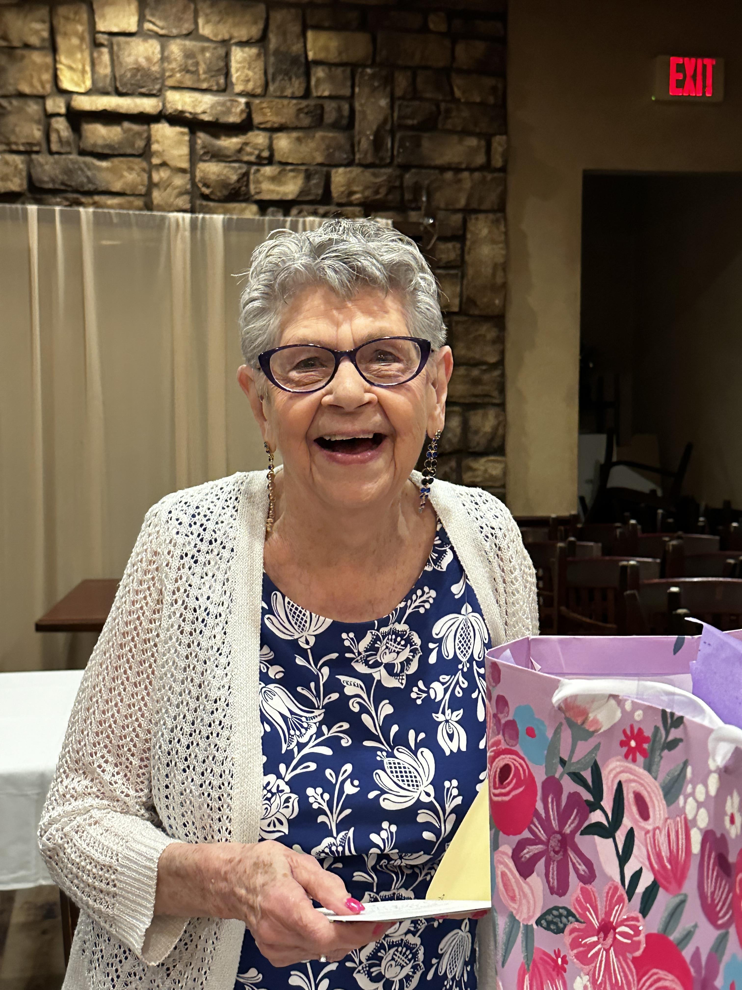 An elderly woman joyfully holds a gift bag while celebrating with friends in a warm setting.