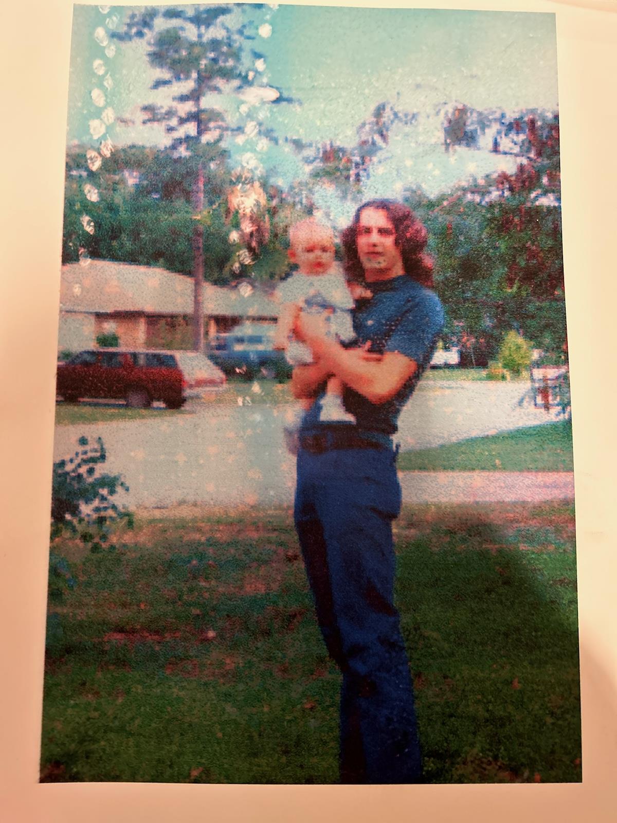 A man holds a baby while standing outside on a grassy area in a residential setting.
