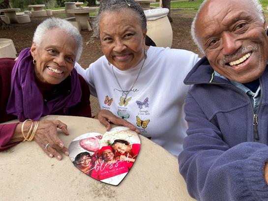 Three friends smile while holding a heart-shaped card in a park on a bright day.