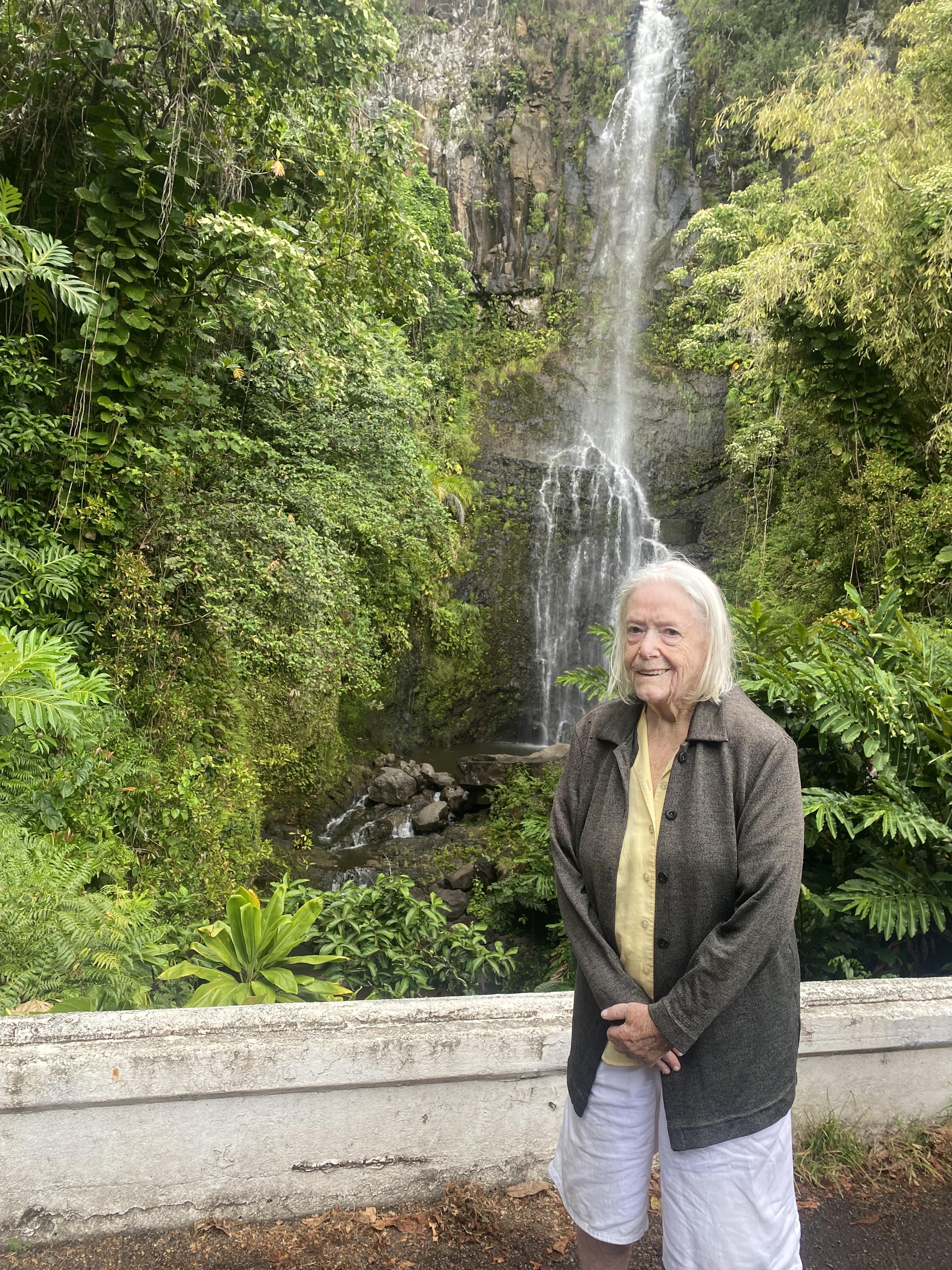 An elderly woman stands gracefully by a stunning waterfall in a lush tropical forest.